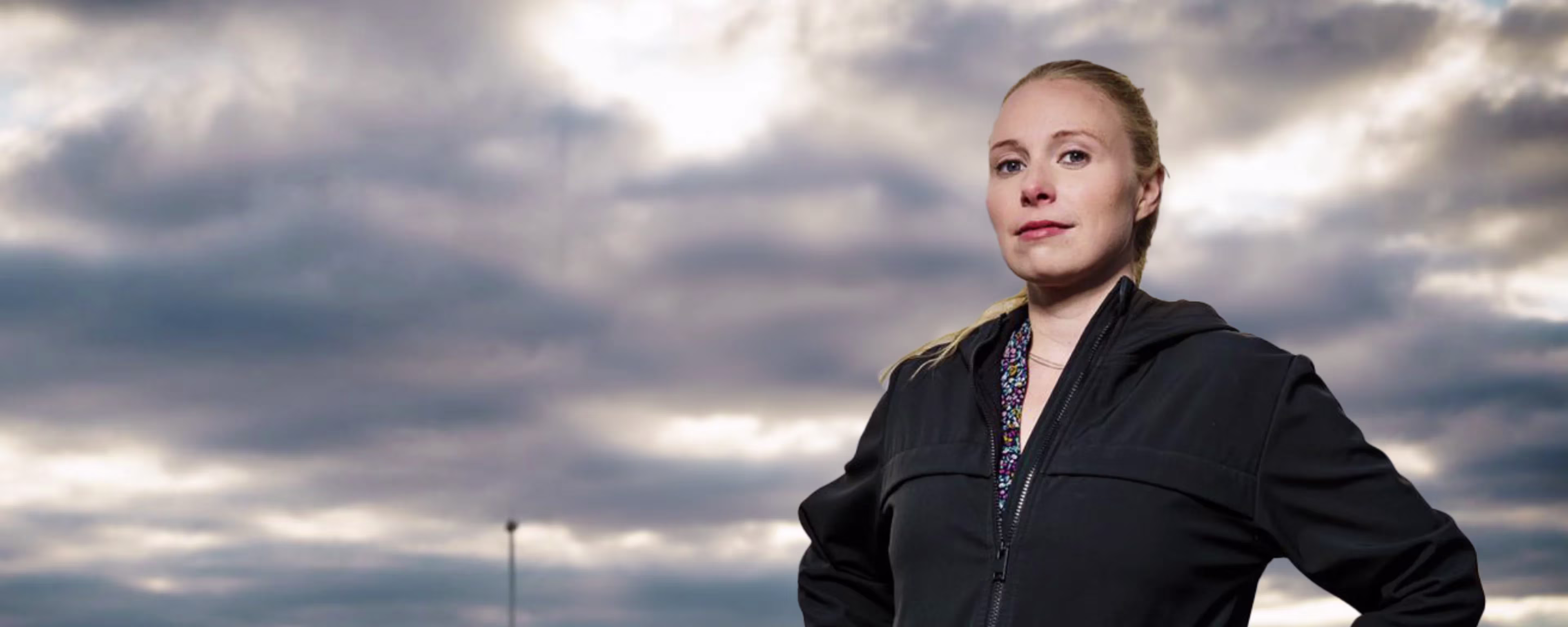 Een vrouw in zwarte jas staat zelfverzekerd voor een bewolkte hemel, handen in de zij, als portret voor SIPOR onderwijs.