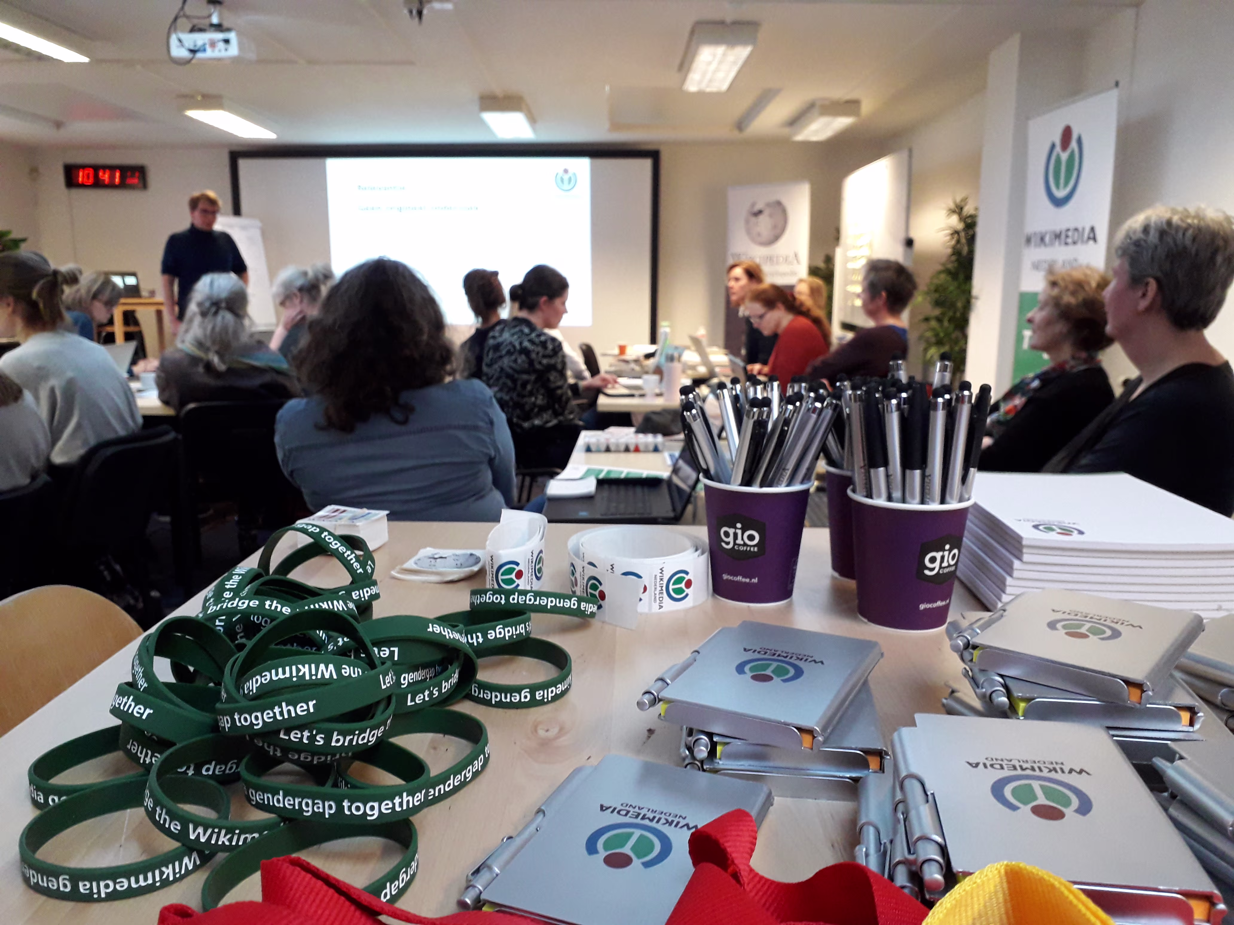 Een presentatie in een kantoorruimte met medewerkers aan tafels, vooraan liggen Wikimedia-materialen zoals lanyards en folders.