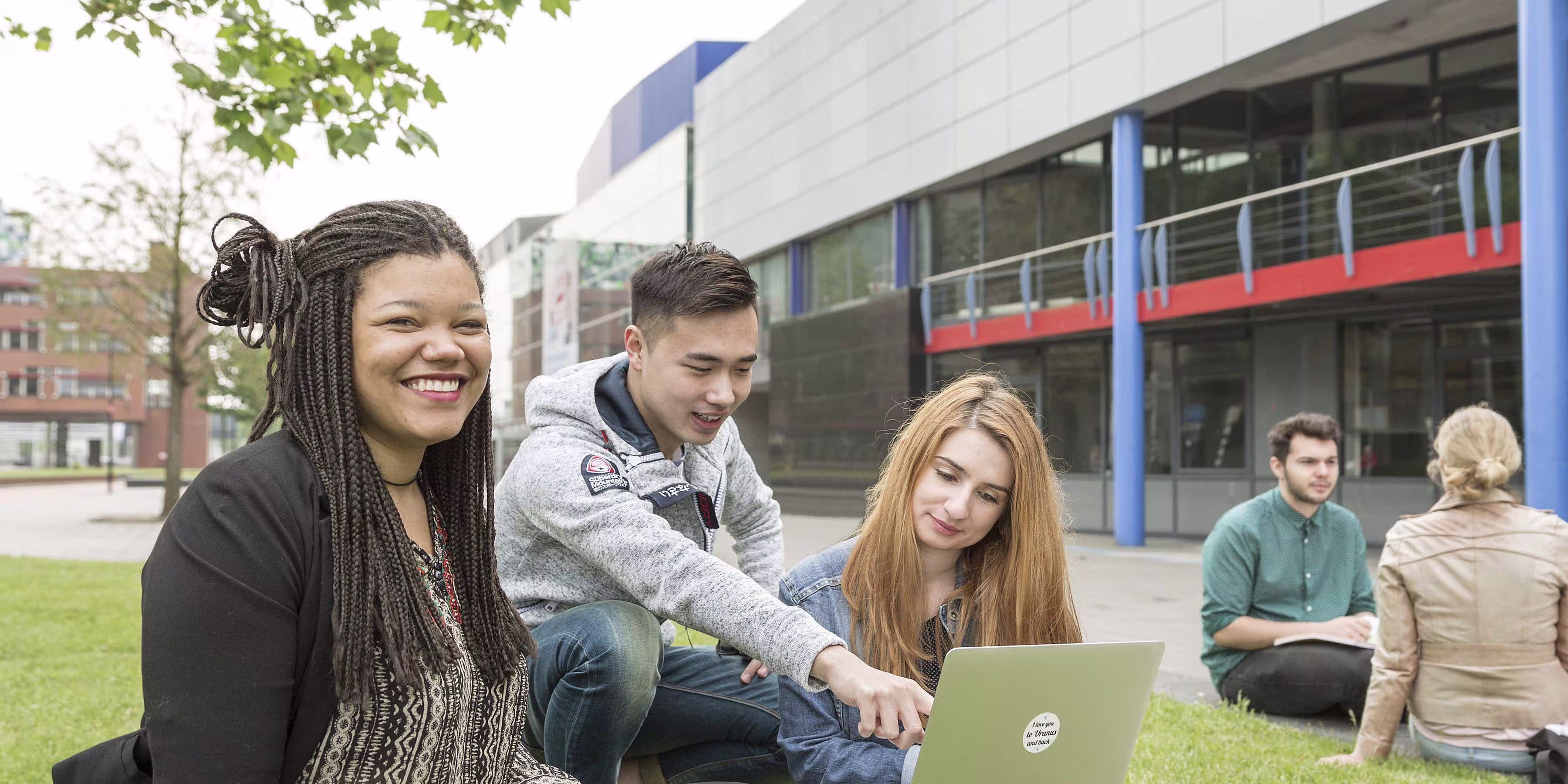 Drie diverse studenten zitten samen op het gras met een laptop voor een modern universiteitsgebouw.
