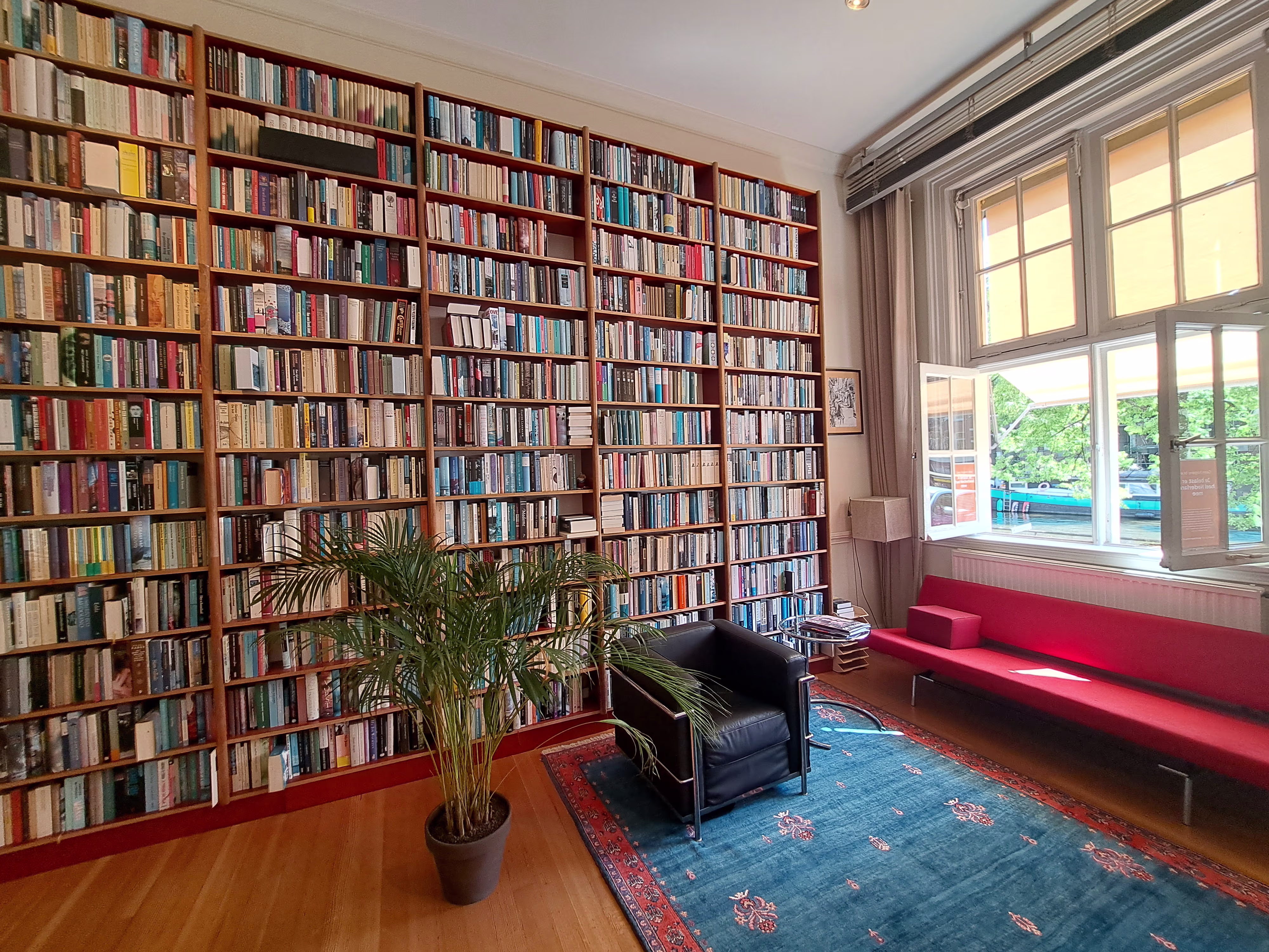 Een gezellige bibliotheek met een enorme boekenwand, zwarte fauteuil, rode bank en grote ramen met uitzicht op groen.