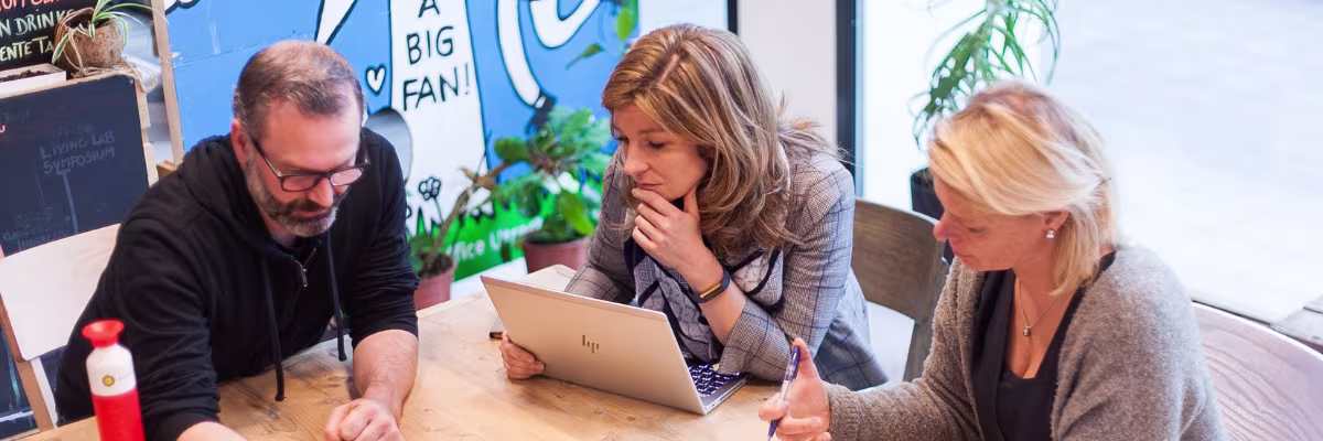Drie collega's werken samen aan een laptop in een moderne kantooromgeving met planten en posters op de achtergrond.