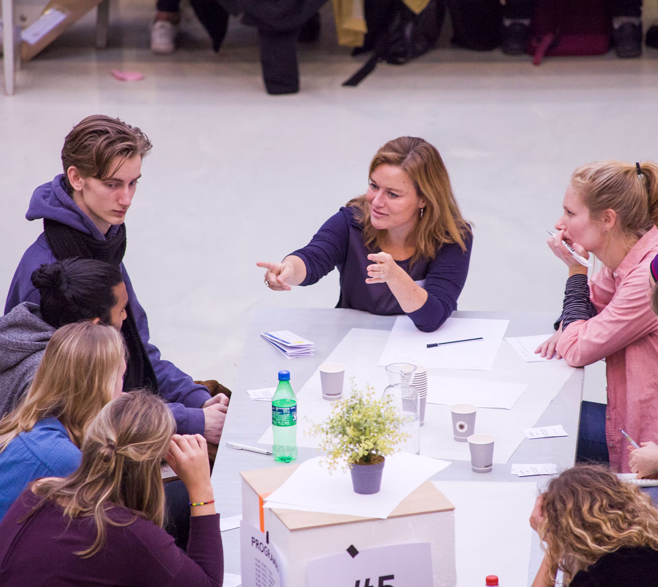 Groep studenten en docent in gesprek rond tafel tijdens bijeenkomst op HKU, met drankjes en materialen op tafel.