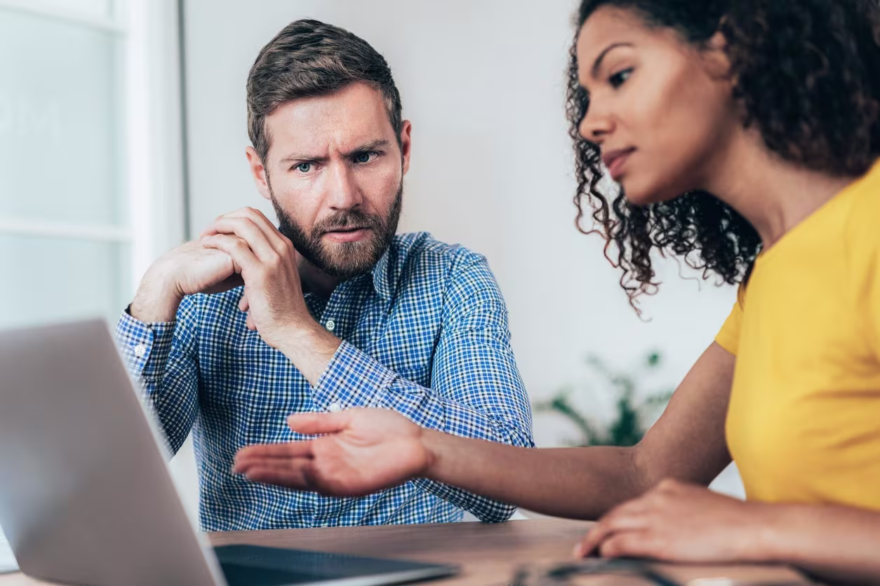 Een man en vrouw in gesprek achter een laptop, de man kijkt peinzend terwijl de vrouw iets uitlegt.