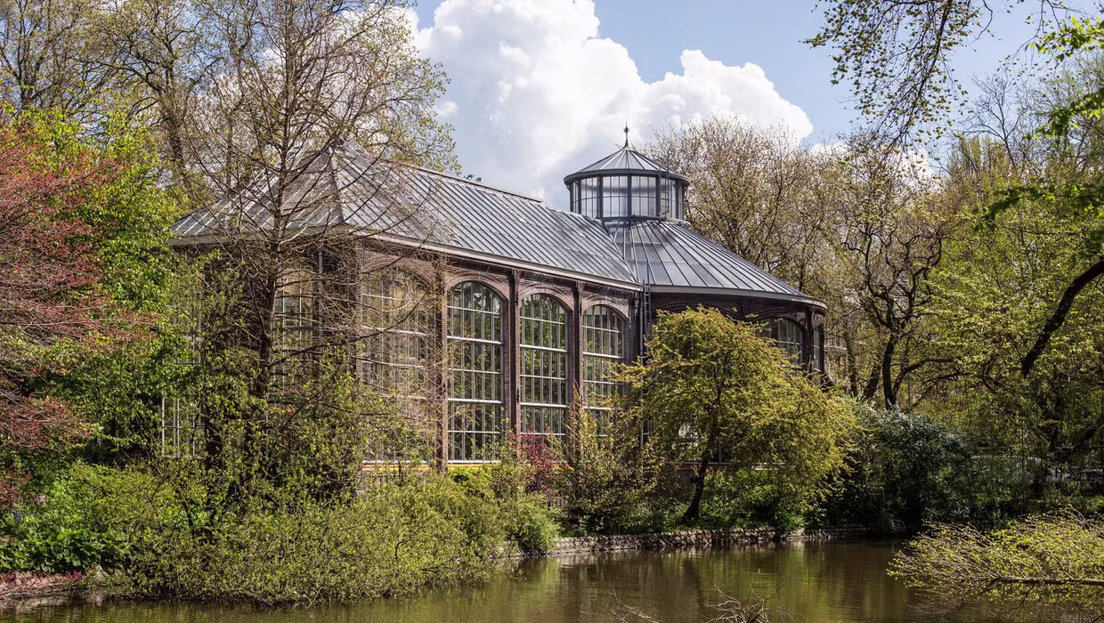 Historische kas met glazen gevel en koepel aan het water, omgeven door groen in de Hortus Botanicus Amsterdam.