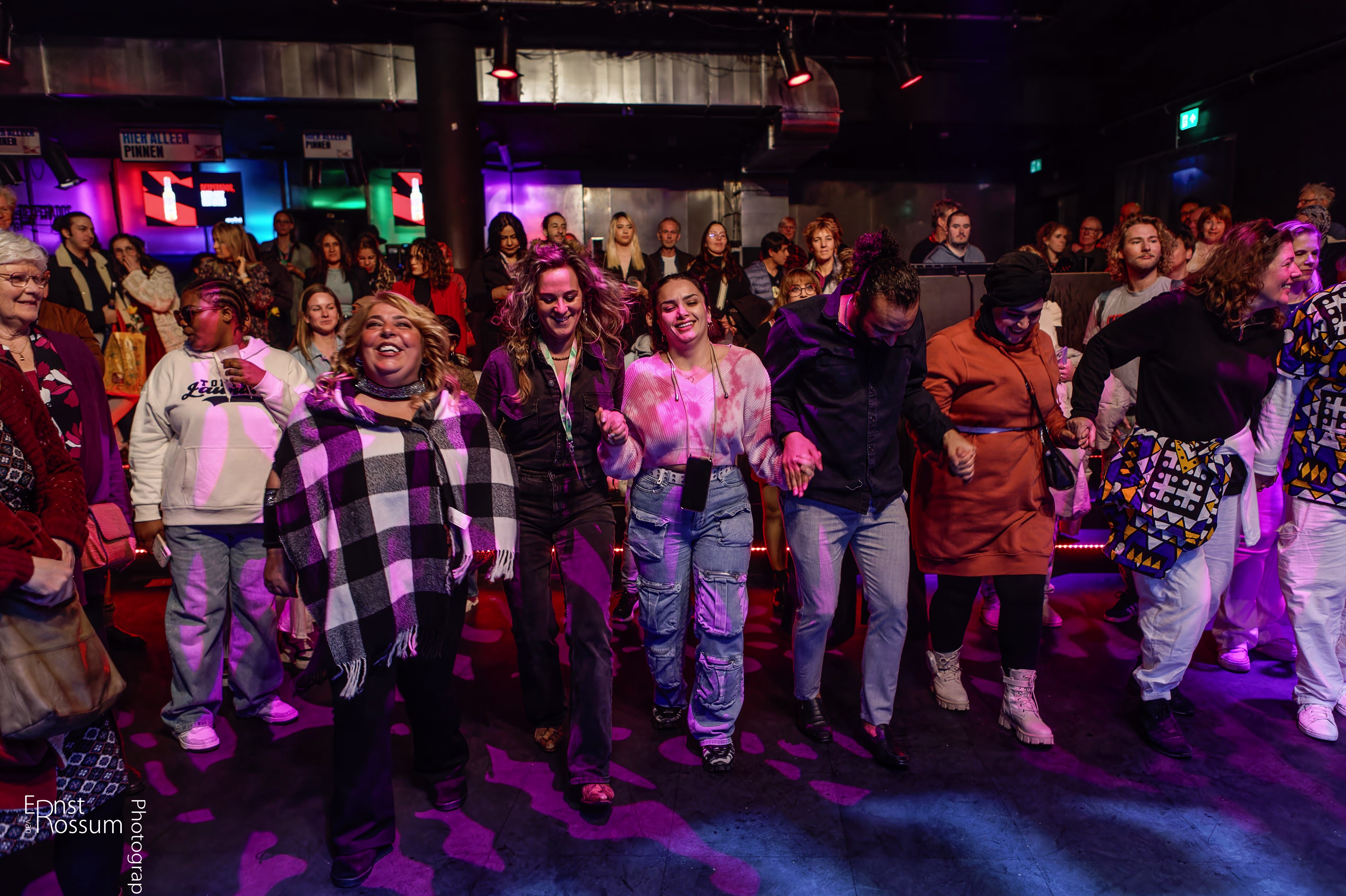 Groep mensen danst samen op een feest in een theater of culturele ruimte met kleurrijke verlichting en vrolijke sfeer.