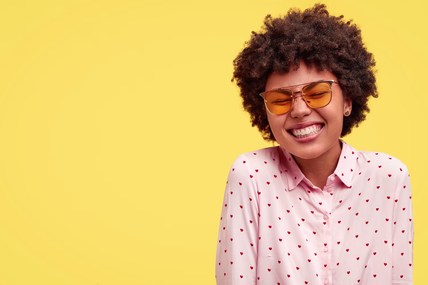 Vrolijke vrouw met krullend haar en gele zonnebril lacht breed, draagt roze shirt met hartjesprint tegen gele achtergrond.