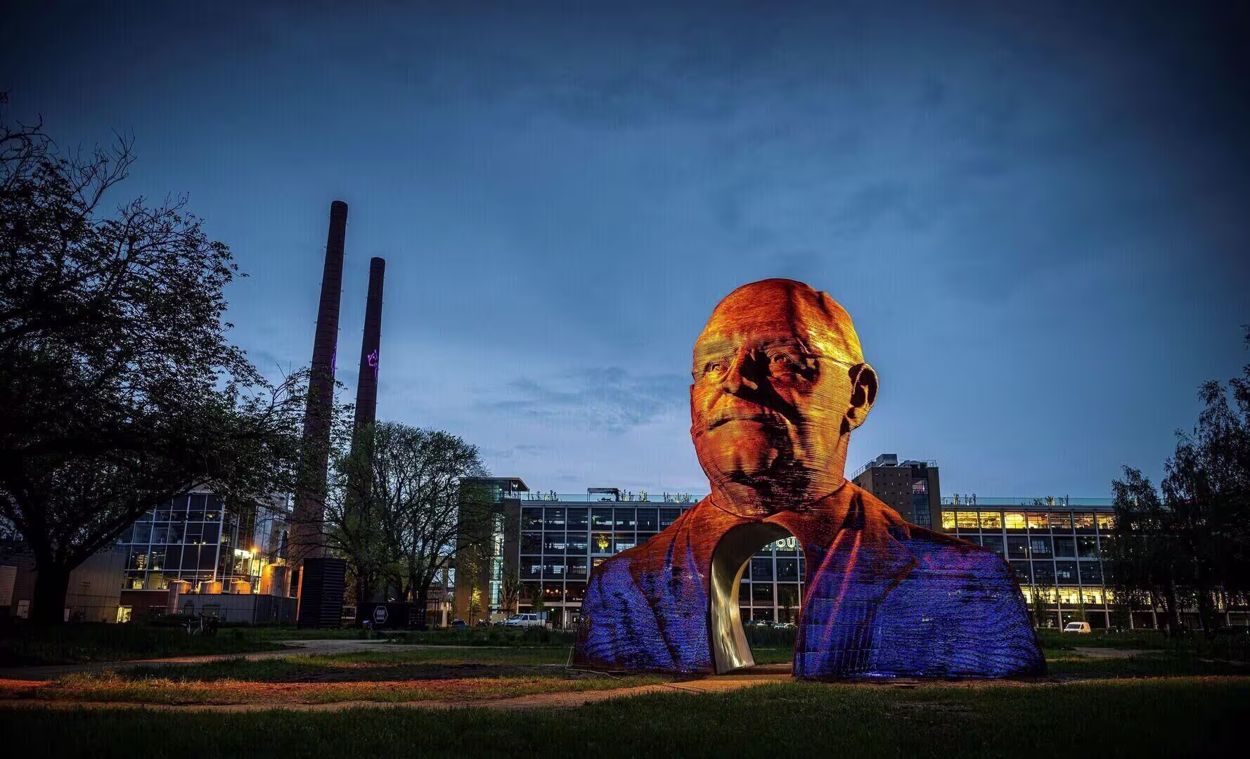 Een groot verlicht kunstwerk van een hoofd staat op een grasveld bij moderne gebouwen in de schemering.