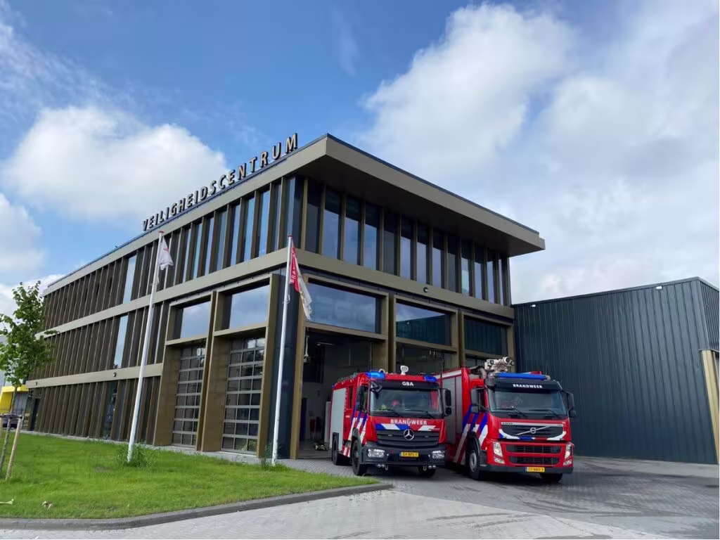 Moderne brandweerkazerne met twee rode brandweerwagens voor de ingang, gebouw heeft glazen gevel en tekst "Veiligheidscentrum".