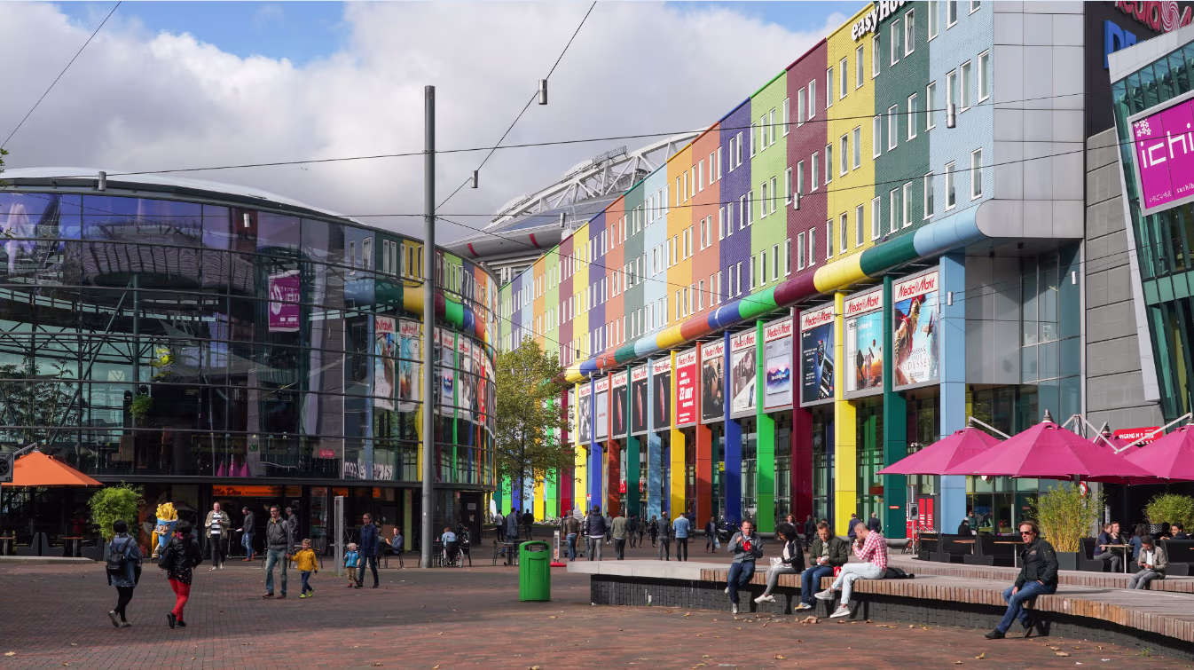 Kleurrijk winkelcentrum met regenbooggevels en terrasjes waar mensen ontspannen op een zonnige dag in Amsterdam.