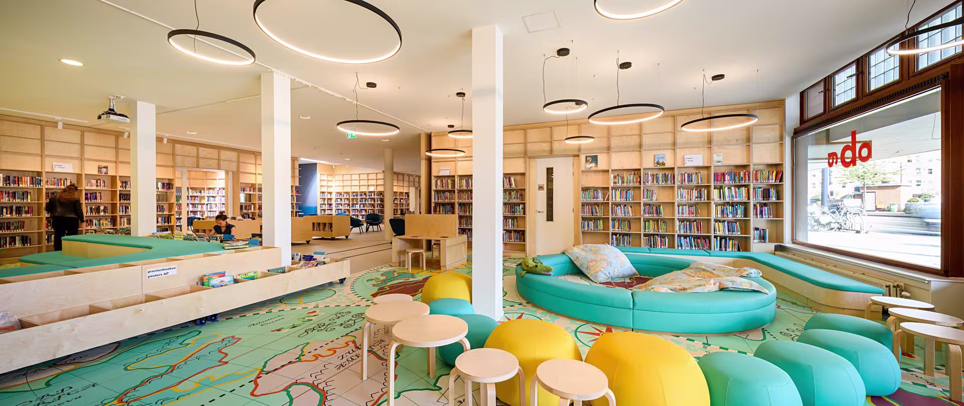 Moderne bibliotheek met kleurrijke zitplekken, boekenkasten en speelse ronde banken in turquoise en geel tinten.