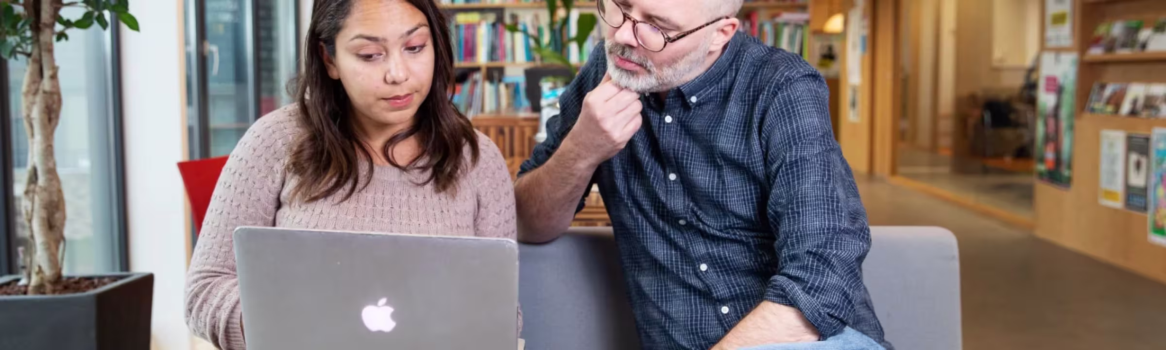 Twee collega's bekijken samen een laptop in een moderne werkruimte met boekenkast op de achtergrond.
