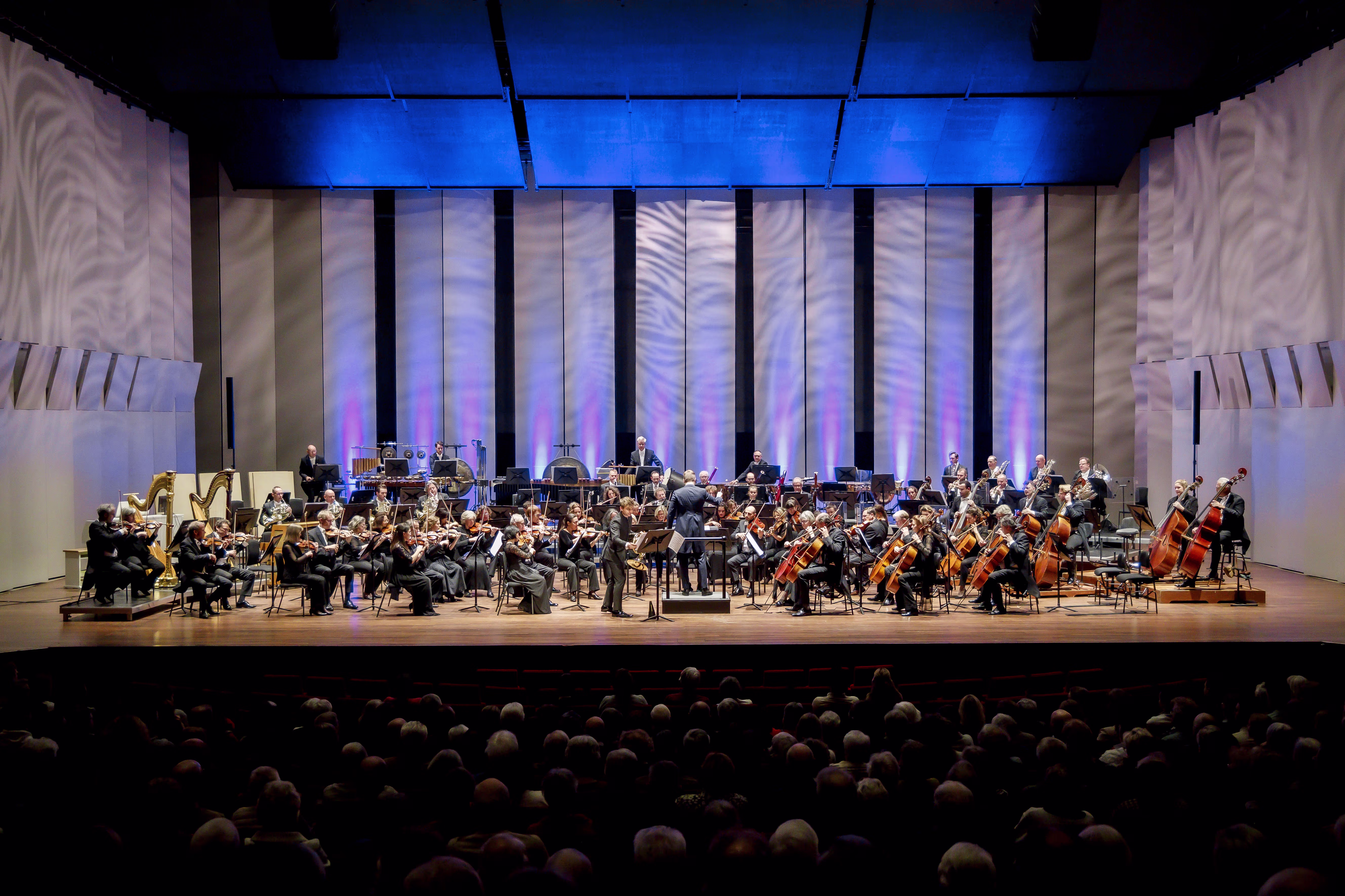 Een symfonieorkest geeft een concert in een moderne concerthal met blauwe verlichting voor een volle zaal publiek.