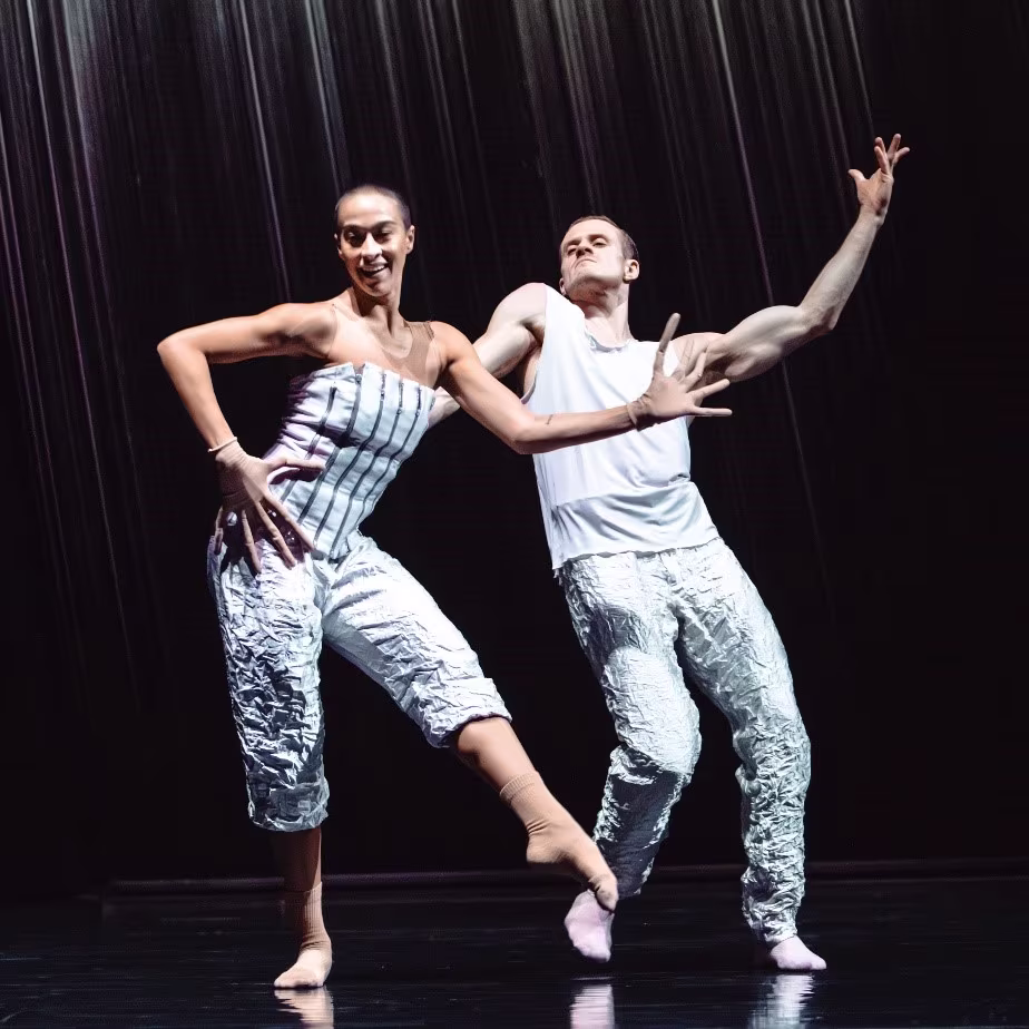 Twee dansers in zilveren outfits voeren een moderne choreografie uit op een donker podium met regeneffect.