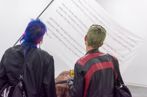 Twee mensen in zwarte jassen bekijken samen tekst op een witte muur in een museum of galerie.