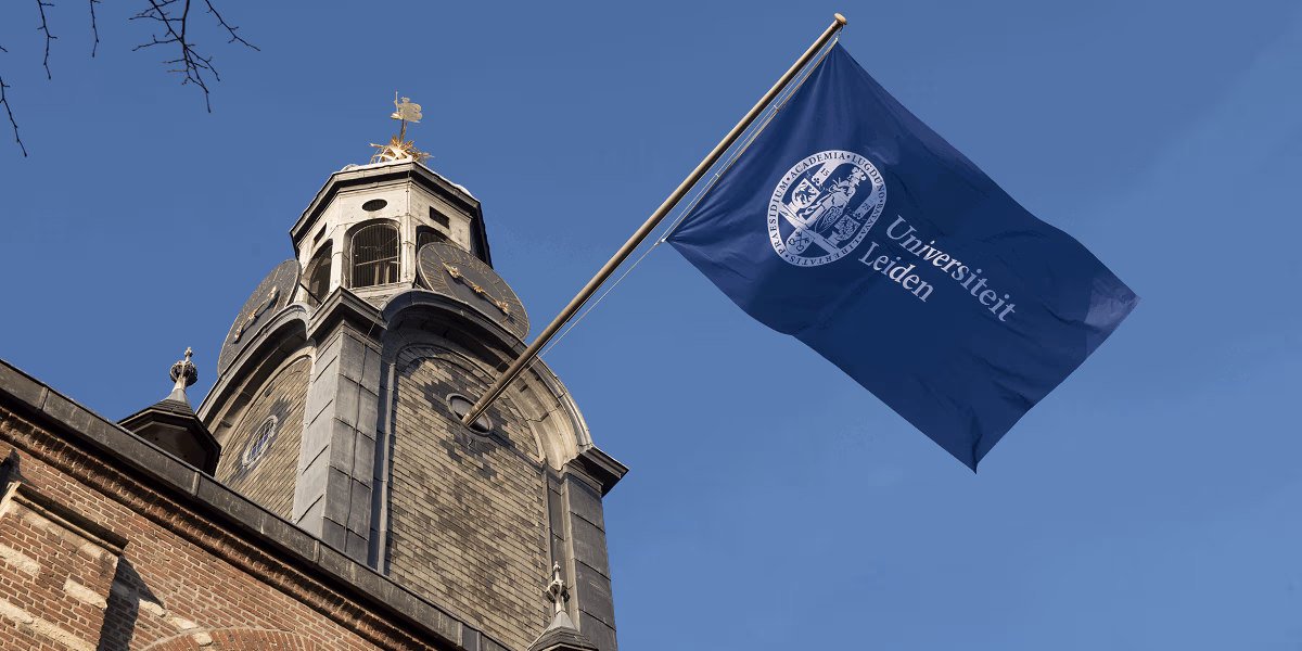 Een historisch gebouw met toren en klok, met daarnaast een blauwe vlag van de Universiteit Leiden wapperend in de wind.