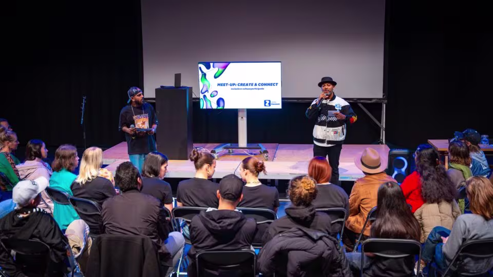 Twee sprekers presenteren op een podium voor een zittend publiek in een theater, met een scherm op de achtergrond.
