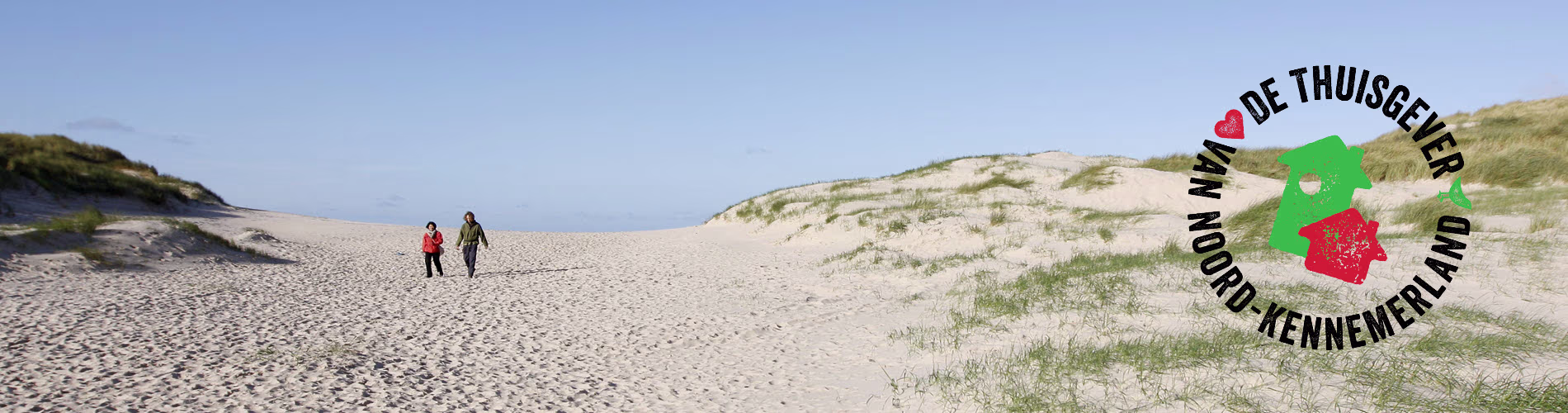Twee personen wandelen over het brede strand van het Noord-Kennemerland met duinen en blauwe lucht op de achtergrond.