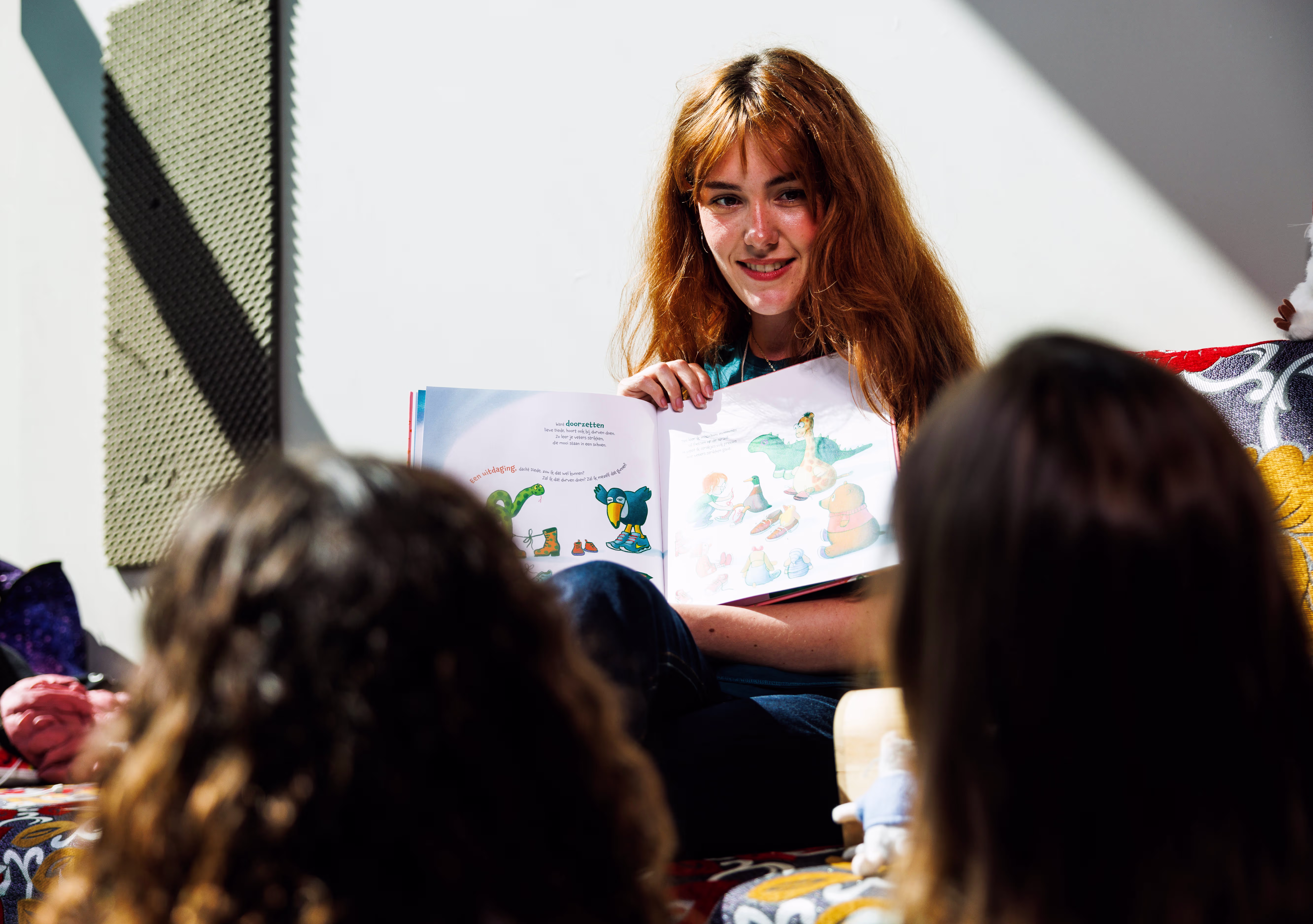 Een vrouw leest voor uit een kinderboek aan een groep kinderen die op de grond zitten te luisteren.