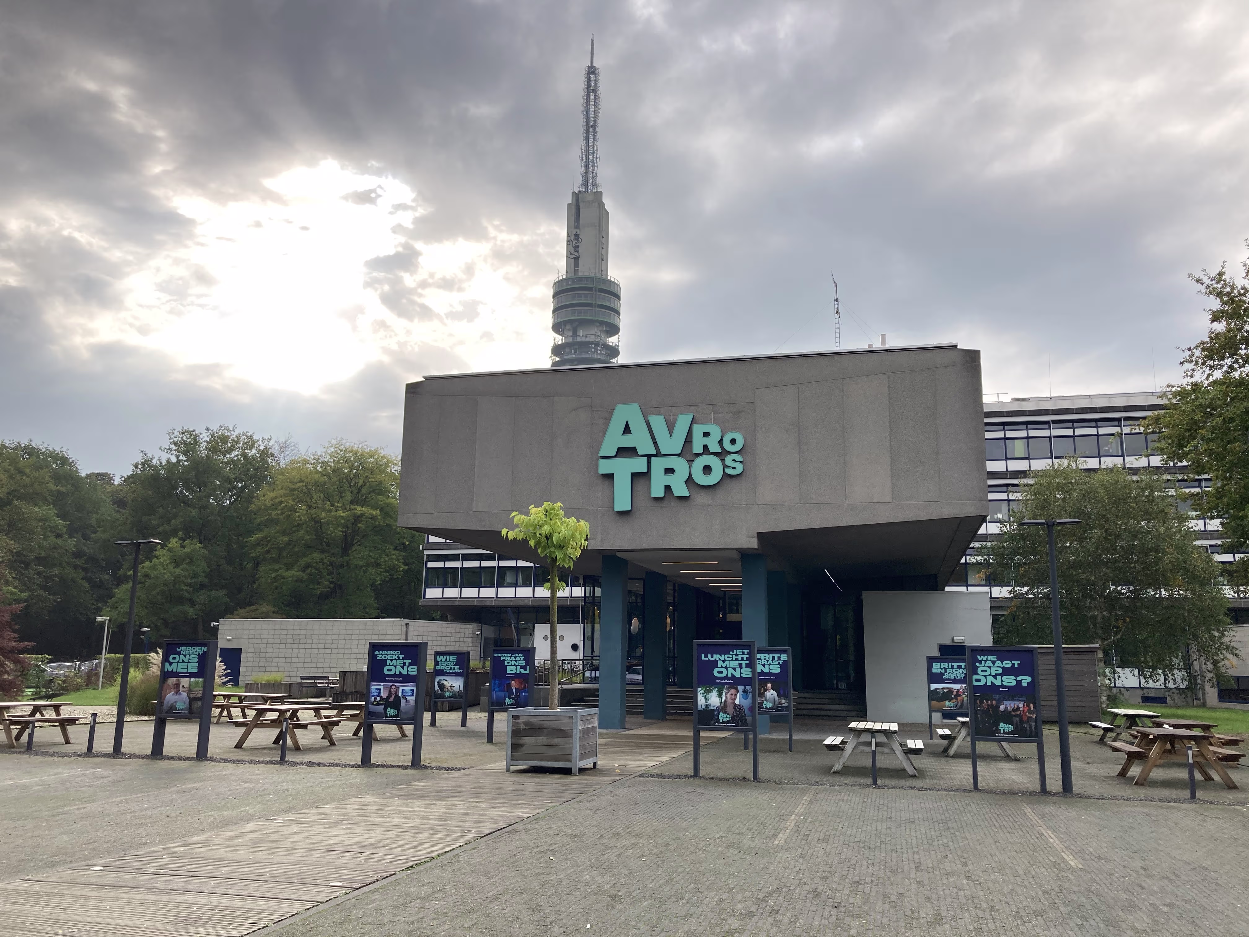 Het AVROTROS-gebouw met logo op de gevel, omgeven door groen en picknicktafels, met een zendmast op de achtergrond.