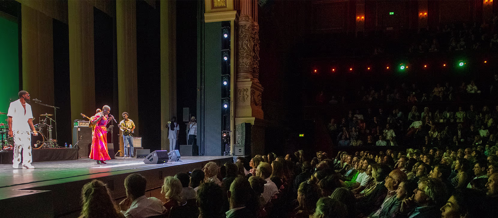Een vol theater kijkt naar een voorstelling op het podium met performers in kleurrijke kostuums onder sfeerverlichting.