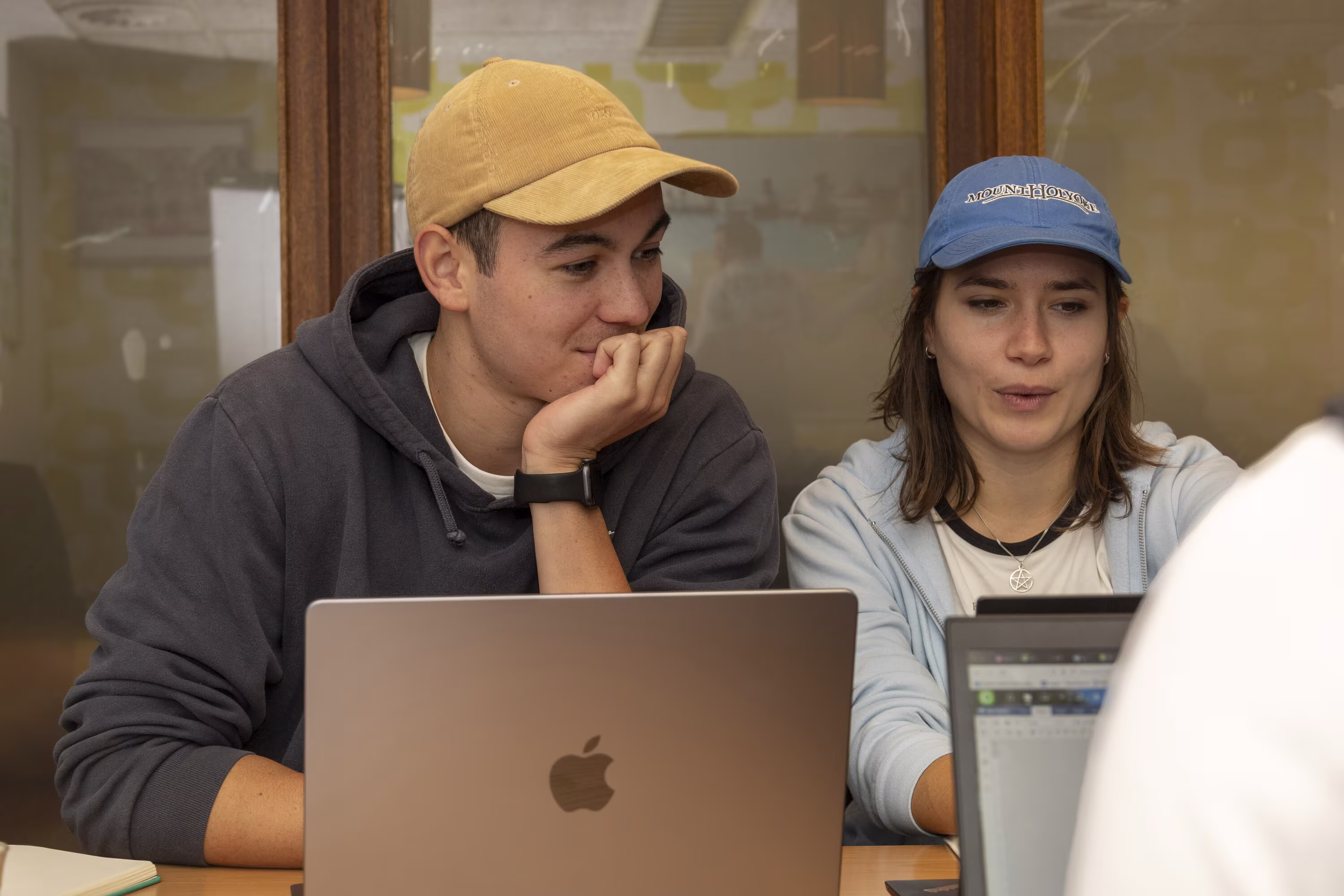 Twee jongeren werken samen achter laptops aan een tafel, beiden dragen petten en kijken geconcentreerd naar hun schermen.