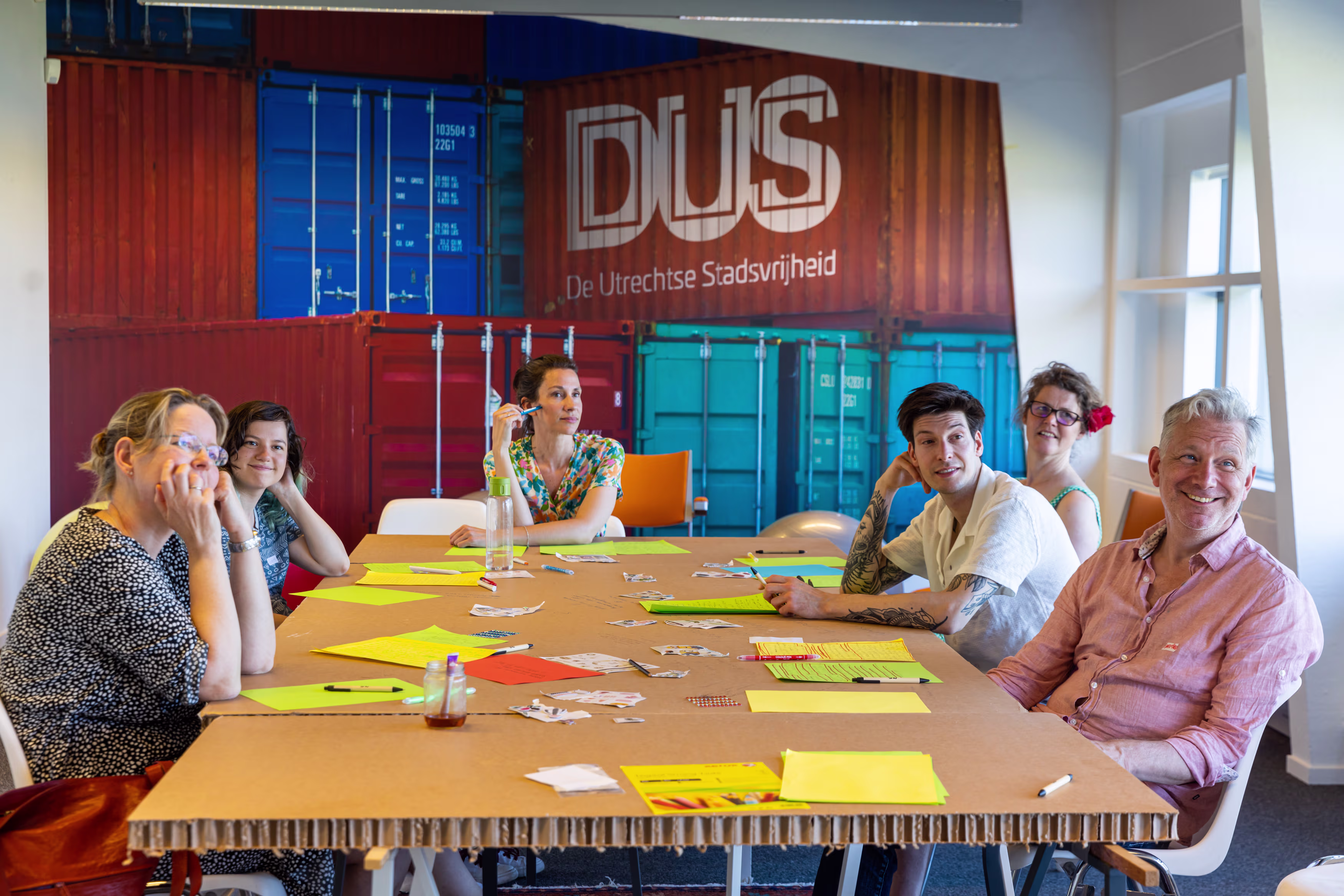 Een groep van zes mensen zit rond een tafel met gele notities tijdens een brainstormsessie in een modern kantoor.