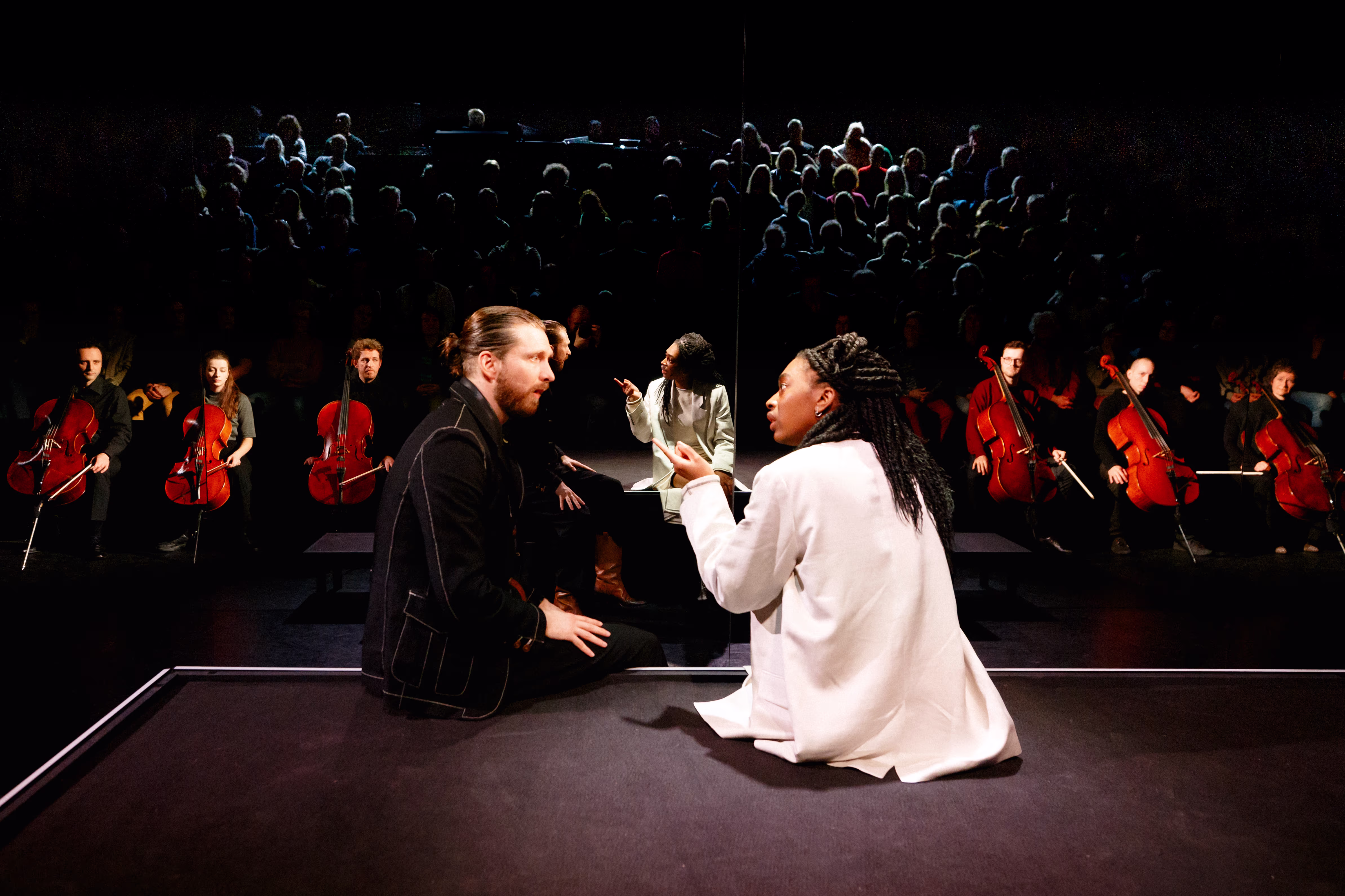 Twee acteurs in een dramatische theaterscène op het podium, omringd door orkestmusici en publiek in een donkere zaal.