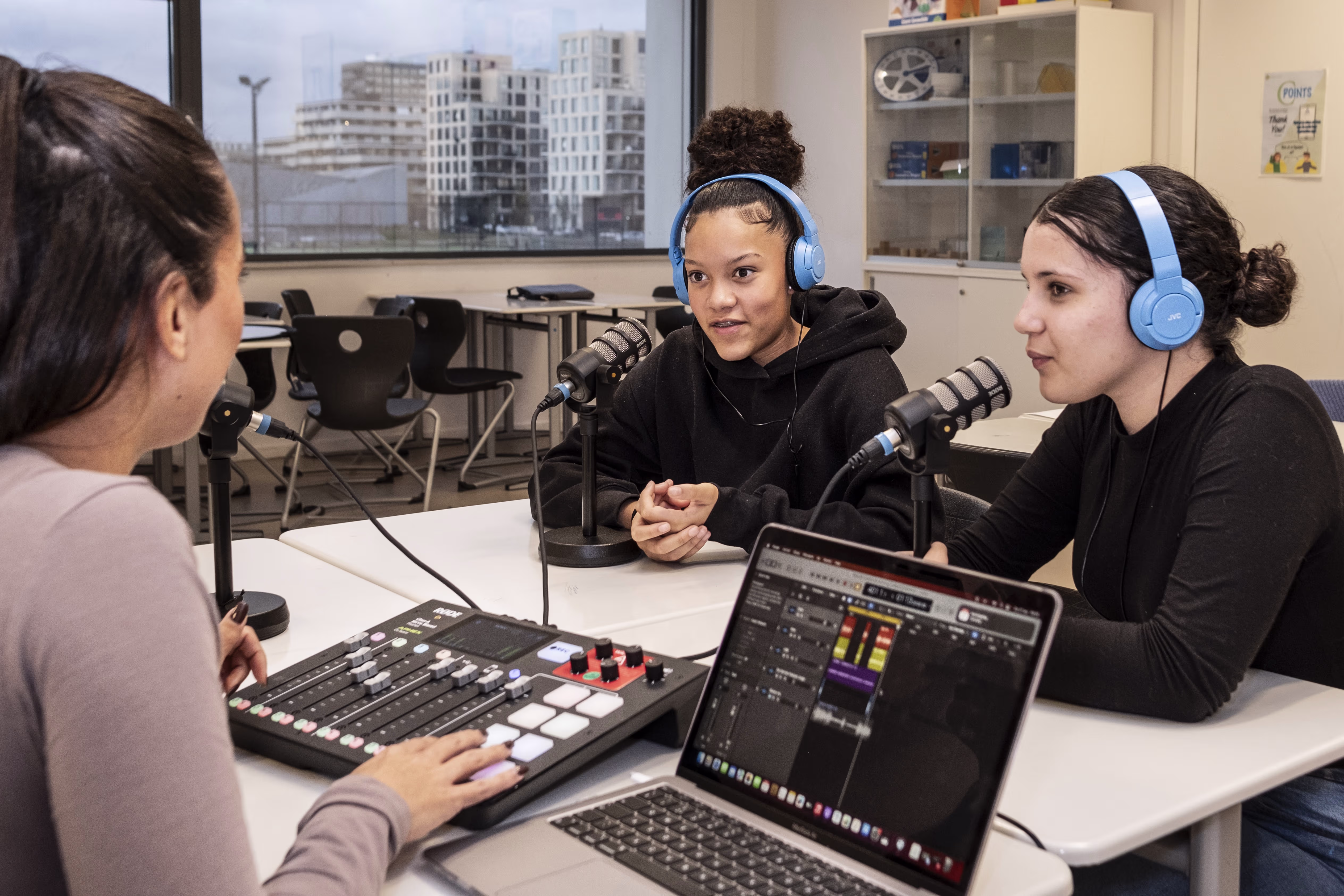 Drie vrouwen in een radiostudio; twee met koptelefoons bij microfoons, één bedient een mengpaneel en laptop.