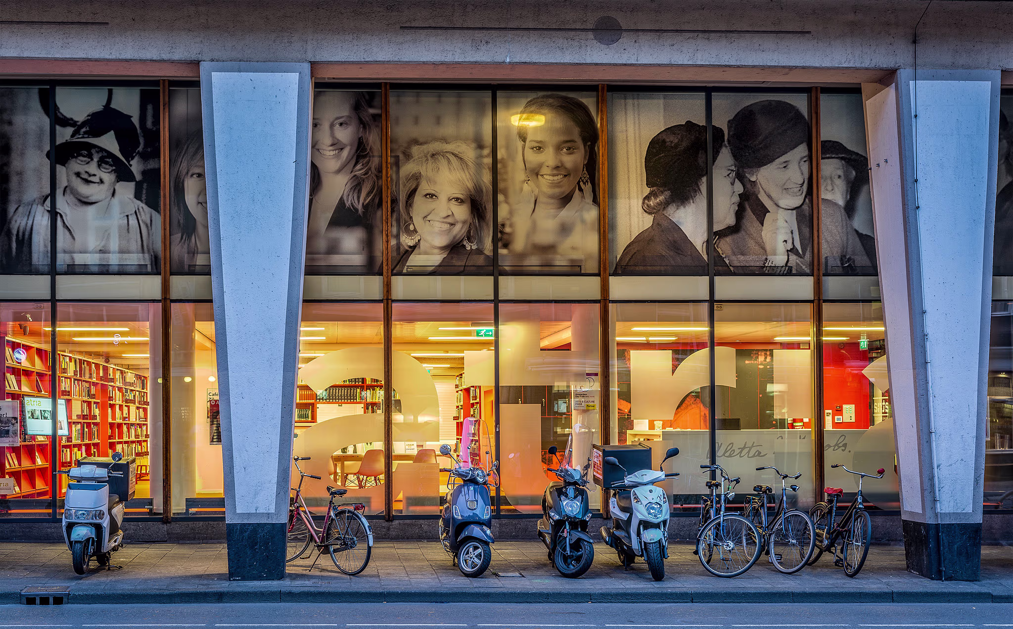 Een bibliotheek met grote ramen waarin portretten van vrouwen zijn getoond, met fietsen en scooters ervoor geparkeerd.