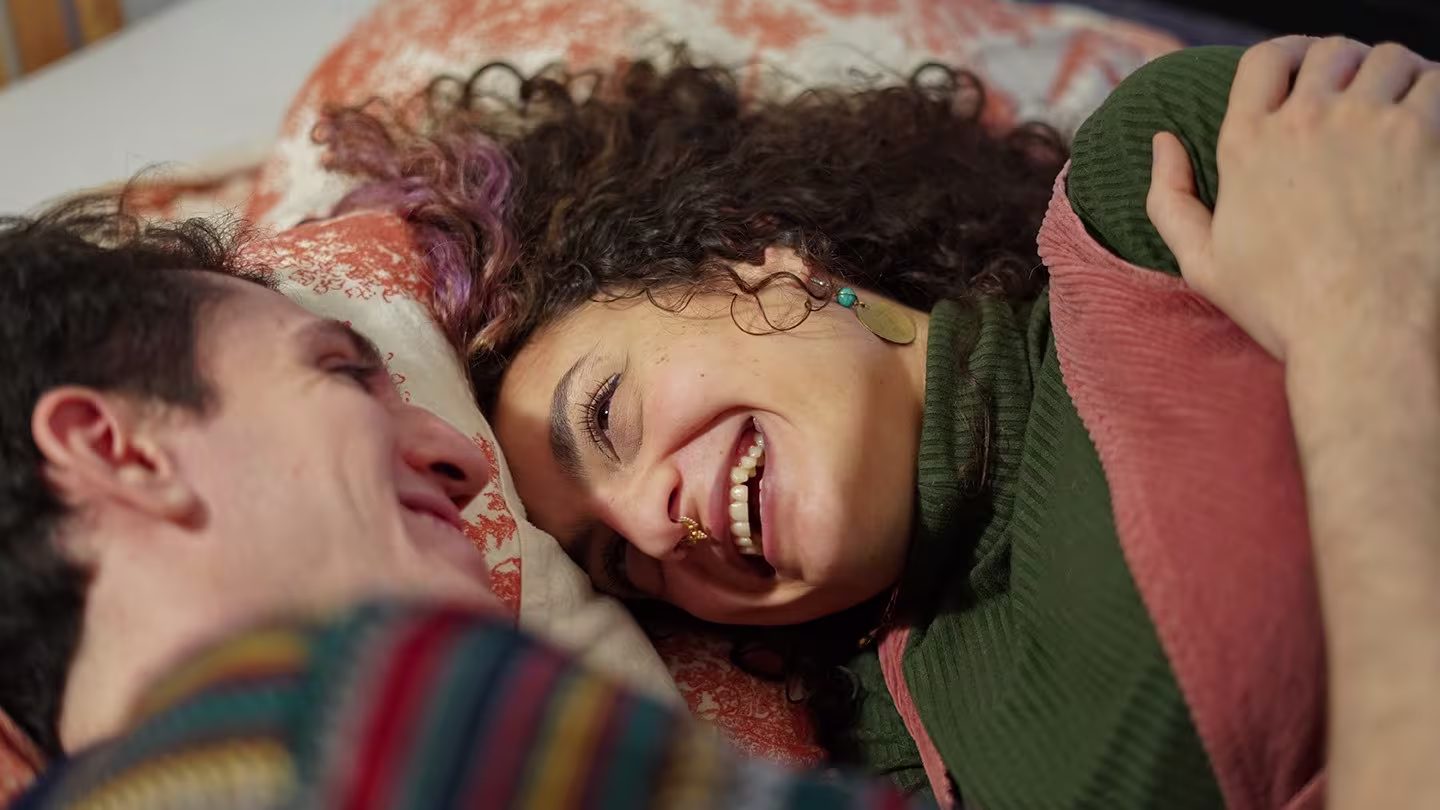 Twee lachende mensen liggen samen ontspannen op bed en kijken elkaar liefdevol aan.