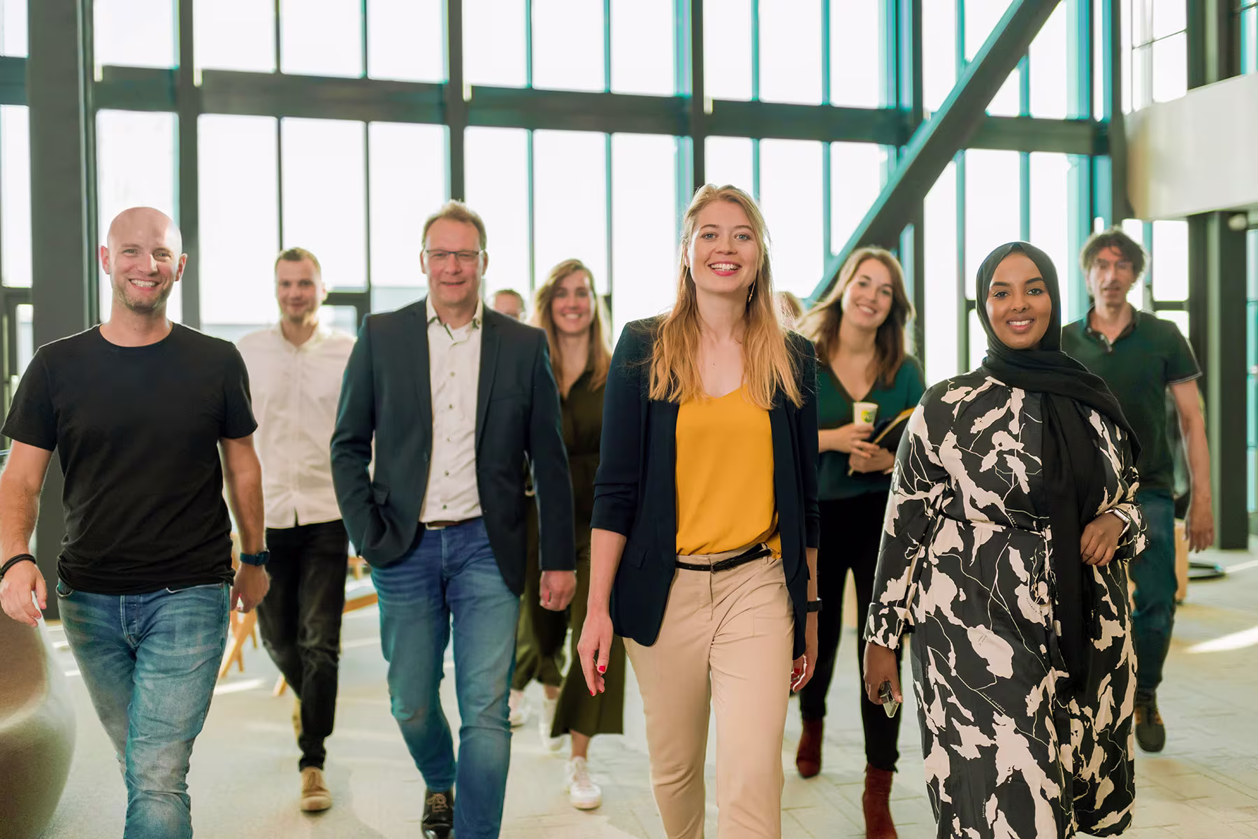 Een diverse groep collega's loopt samen door een moderne kantooromgeving met grote ramen en veel natuurlijk licht.