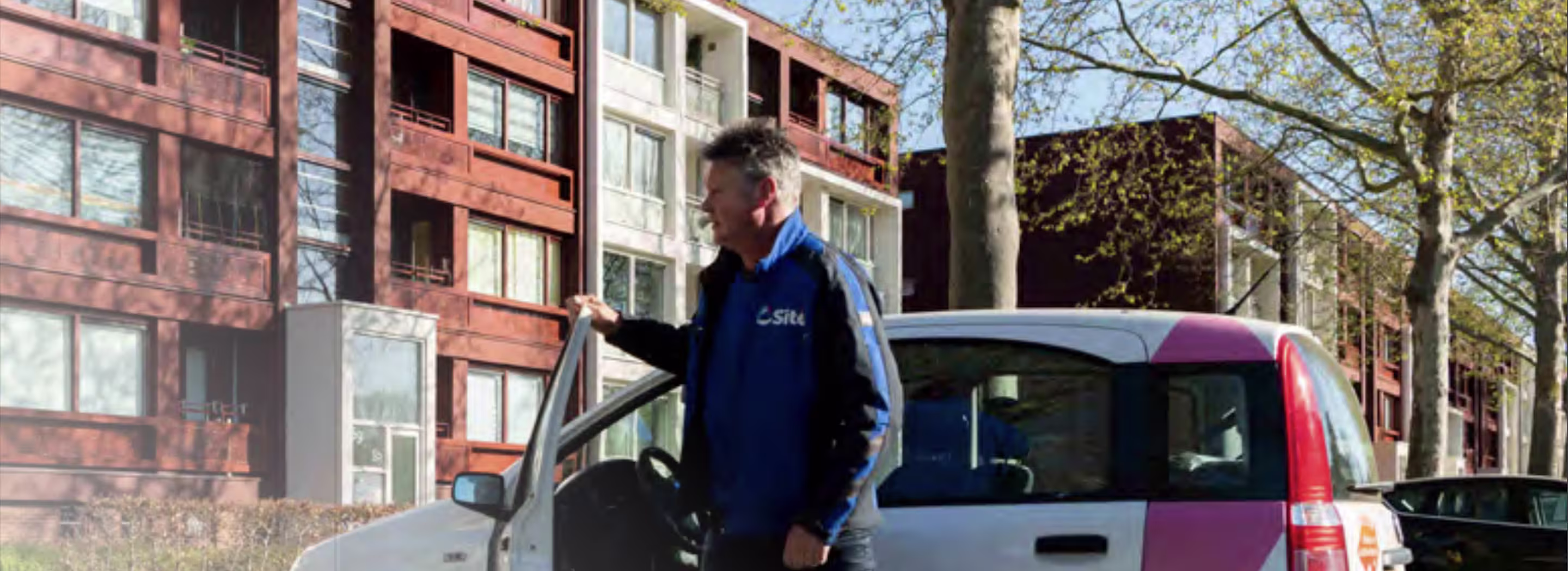 Een medewerker in blauw Sité-jack staat bij een auto voor moderne appartementen.
