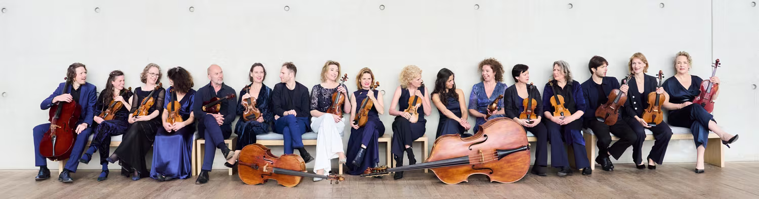 Orkestleden van Amsterdam Sinfonietta zitten in een rij met hun strijkinstrumenten tegen een witte muur.