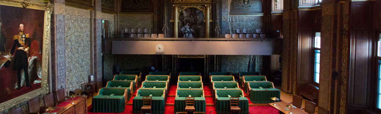 Vergaderzaal van de Eerste Kamer met groene stoelen, rode vloerbedekking en historische portretten aan de muren.