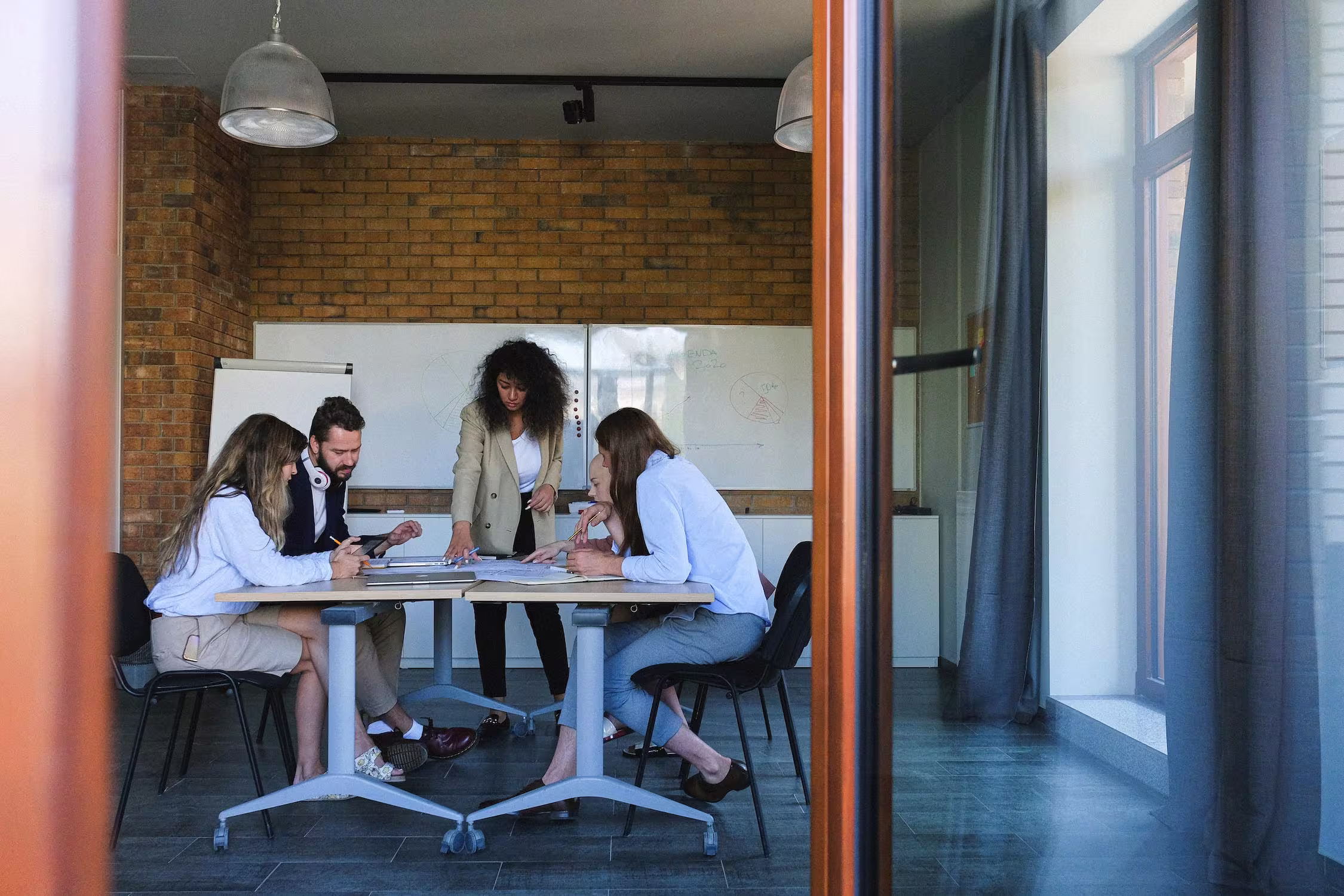 Vier collega's in een moderne vergaderruimte met bakstenen muur bespreken documenten aan een witte tafel.