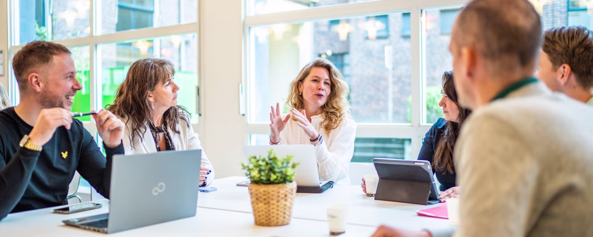 Een groep collega's in vergadering rond een tafel met laptops, waarbij een vrouw in het wit spreekt en gebaart.