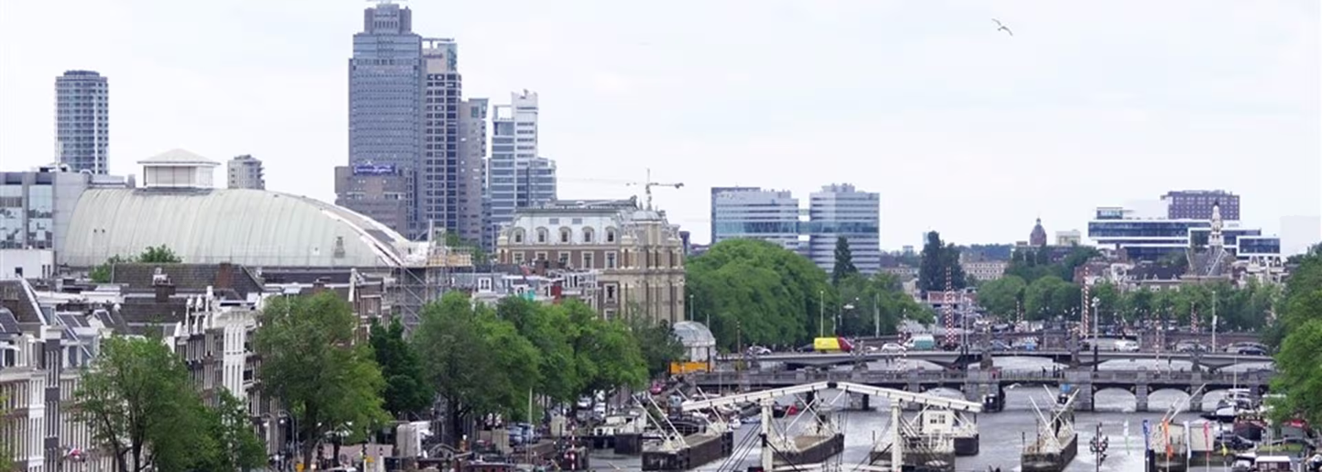Stadsgezicht met moderne kantoorgebouwen, historische architectuur, groene bomen en een gracht met boten en bruggen.