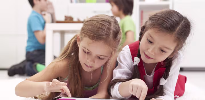 Twee meisjes liggen op de grond en kijken samen naar een tablet, terwijl andere kinderen op de achtergrond spelen.