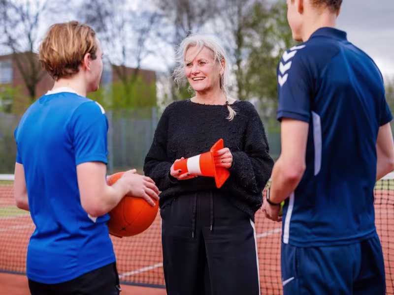 Een vrouw in zwarte kleding praat met twee jongens in blauwe sportkleding op een sportterrein met oranje pionnen.