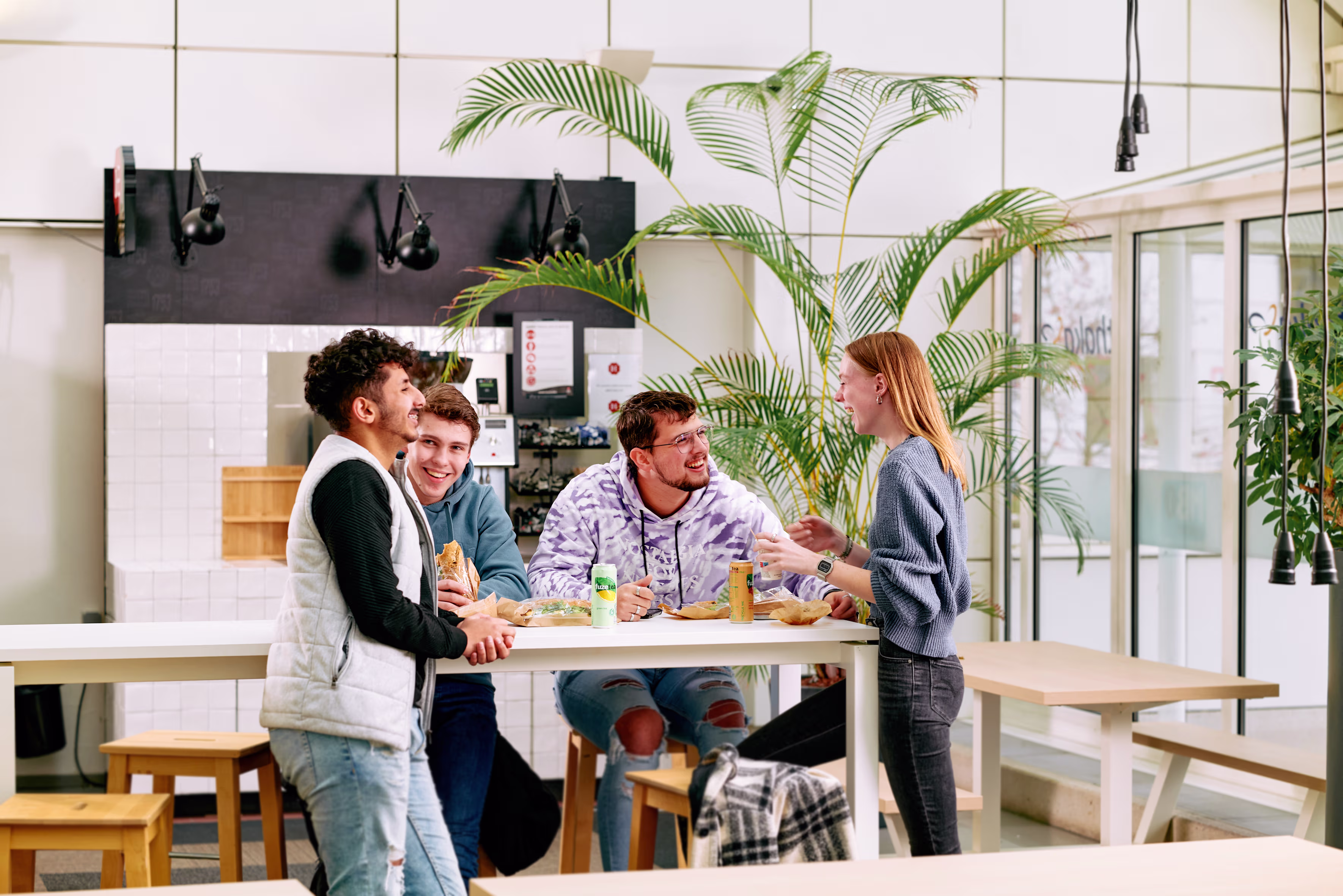 Vier jonge collega's staan lachend rond een tafel in een moderne kantineruimte met planten en natuurlijk licht.