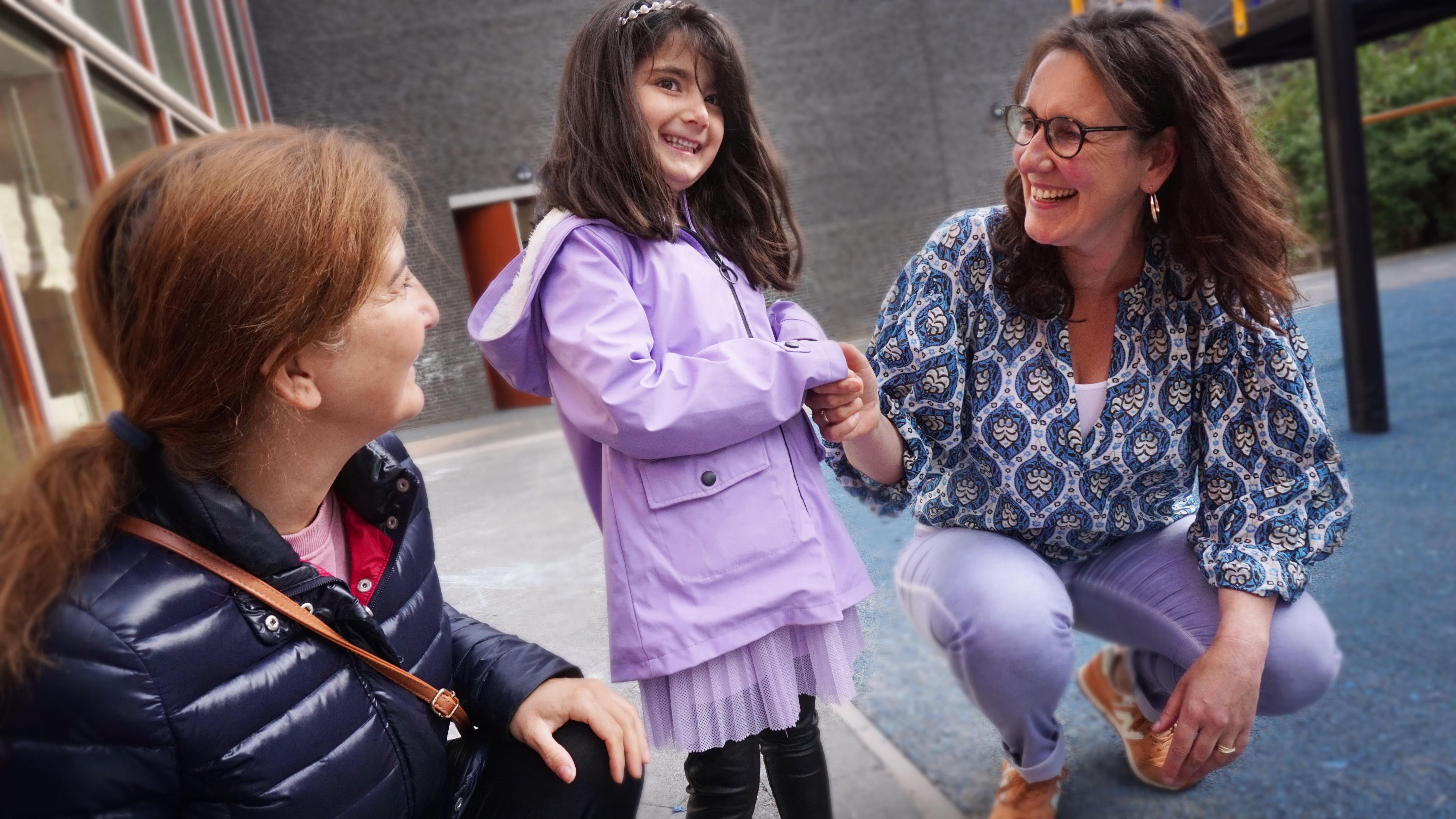 Een vrouw in gesprek met twee meisjes op een schoolplein, allemaal lachend in een ontspannen sfeer.