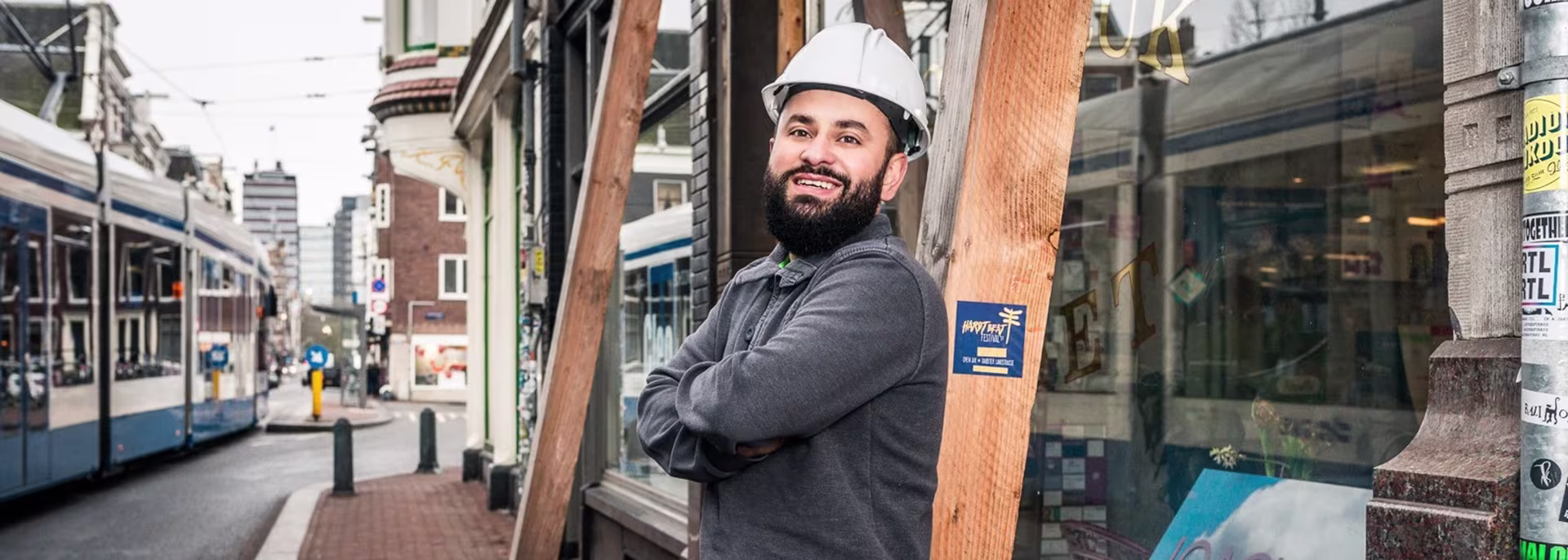 Een lachende man met witte helm en donkere trui staat met gekruiste armen voor een winkelstraat met een tram op de achtergrond.