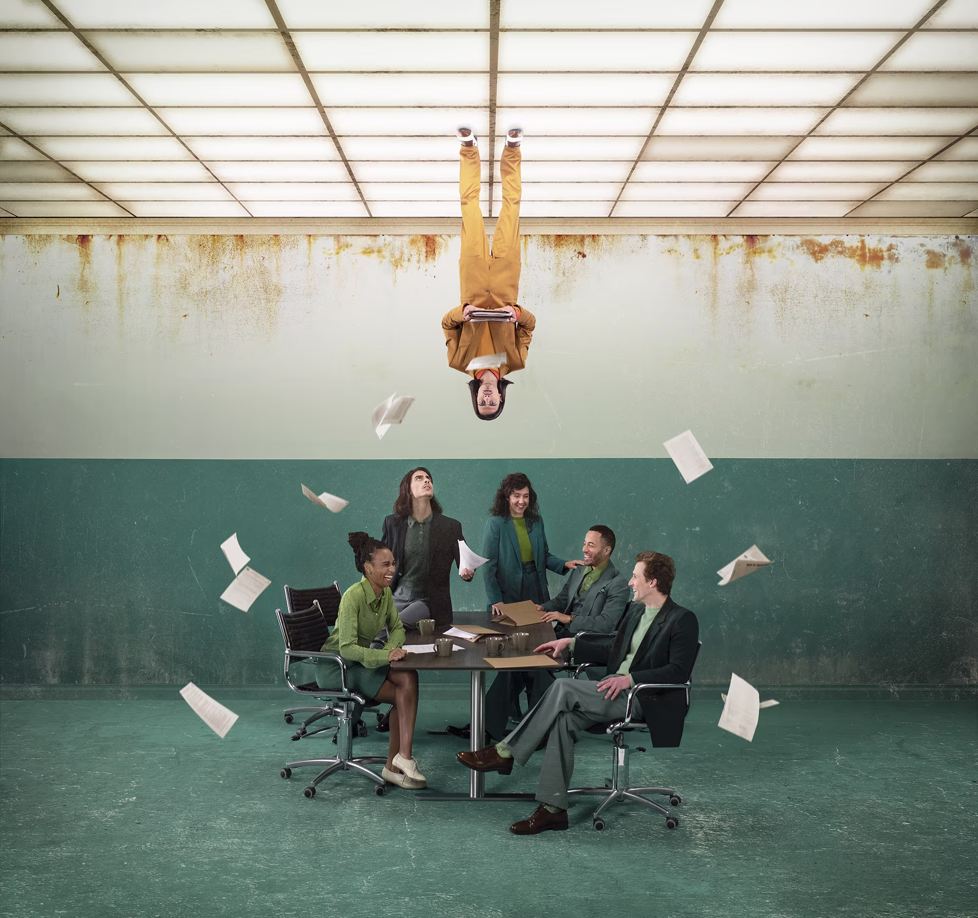 Een groep mensen in pak zit rond een tafel terwijl papieren door de lucht vliegen en één persoon ondersteboven hangt.