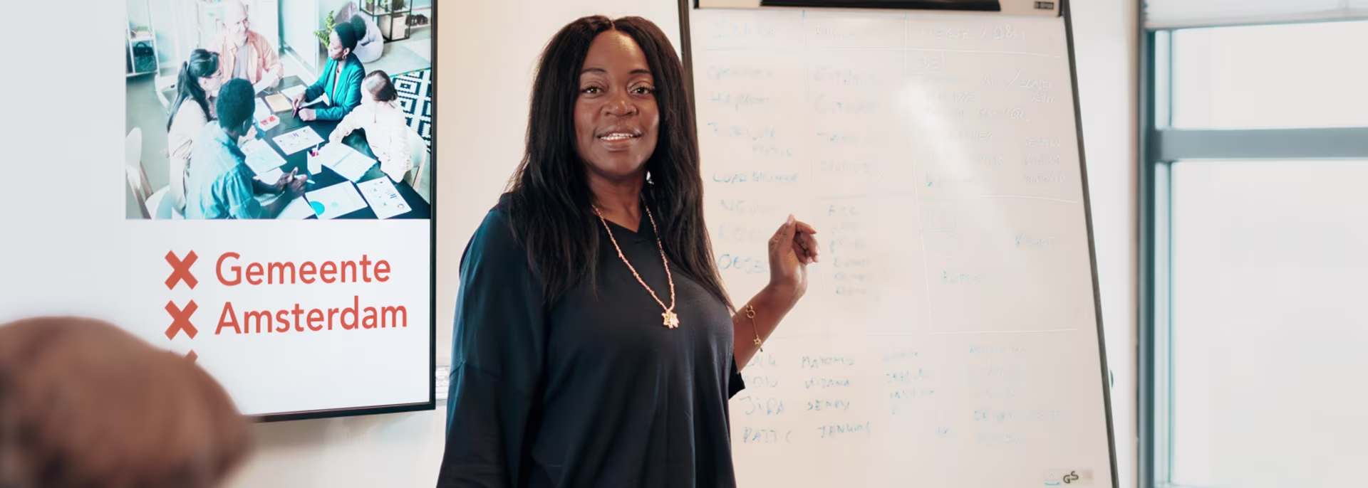 Een vrouw geeft een presentatie voor een whiteboard, met op de achtergrond het logo van Gemeente Amsterdam.