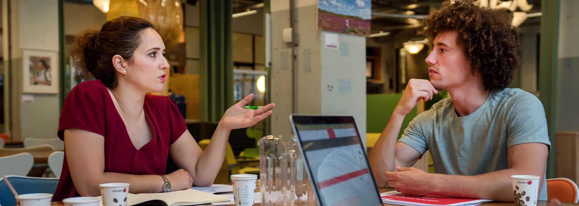 Twee collega's in gesprek aan een tafel met laptop, koffie en water in een modern kantoor van Gemeente Altena.