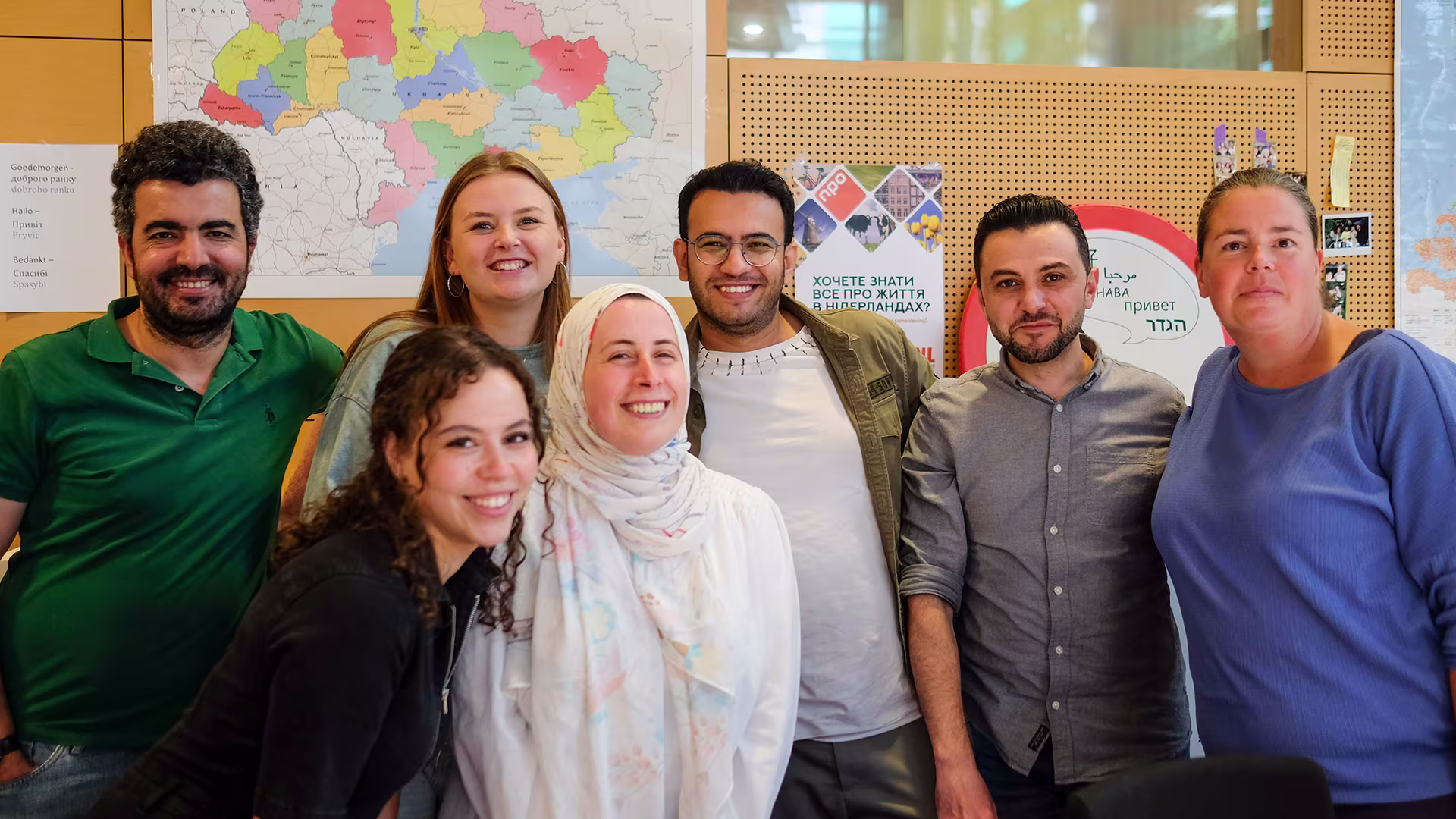 Groepsfoto van zeven lachende collega's in een kantoorruimte met wereldkaart en prikbord op de achtergrond.