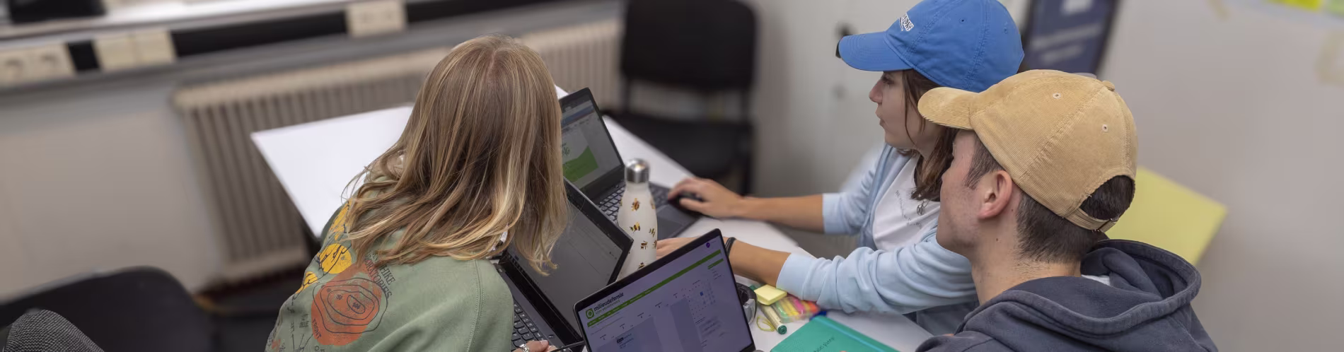 Drie jongeren werken samen achter laptops aan een tafel, een draagt een groene trui, anderen hebben petten op.