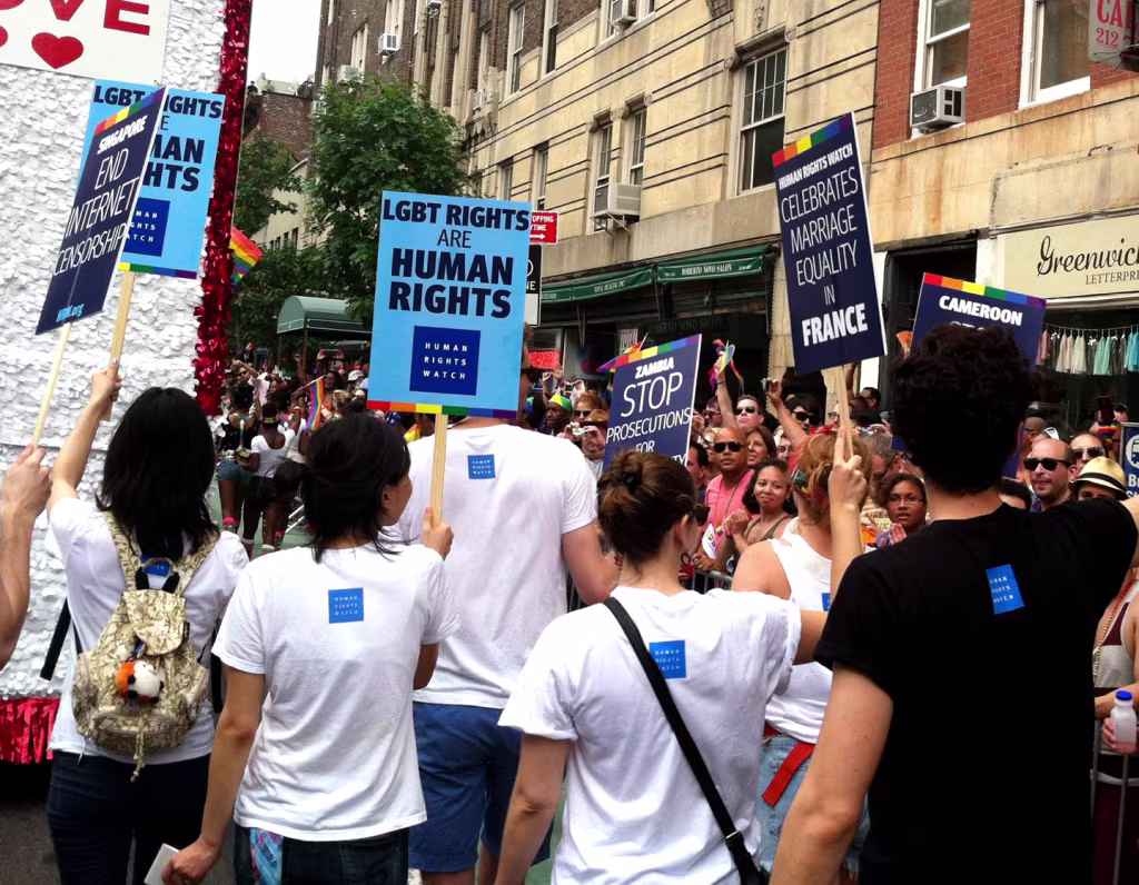 Een menigte demonstranten houdt borden omhoog voor LHBTI-rechten, met teksten zoals "LGBT RIGHTS ARE HUMAN RIGHTS" en "STOP PROSECUTIONS".