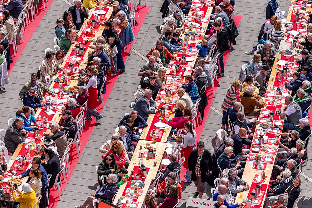 Luchtfoto van mensen die samen aan lange rode tafels zitten tijdens een bevrijdingsmaaltijd op een plein.