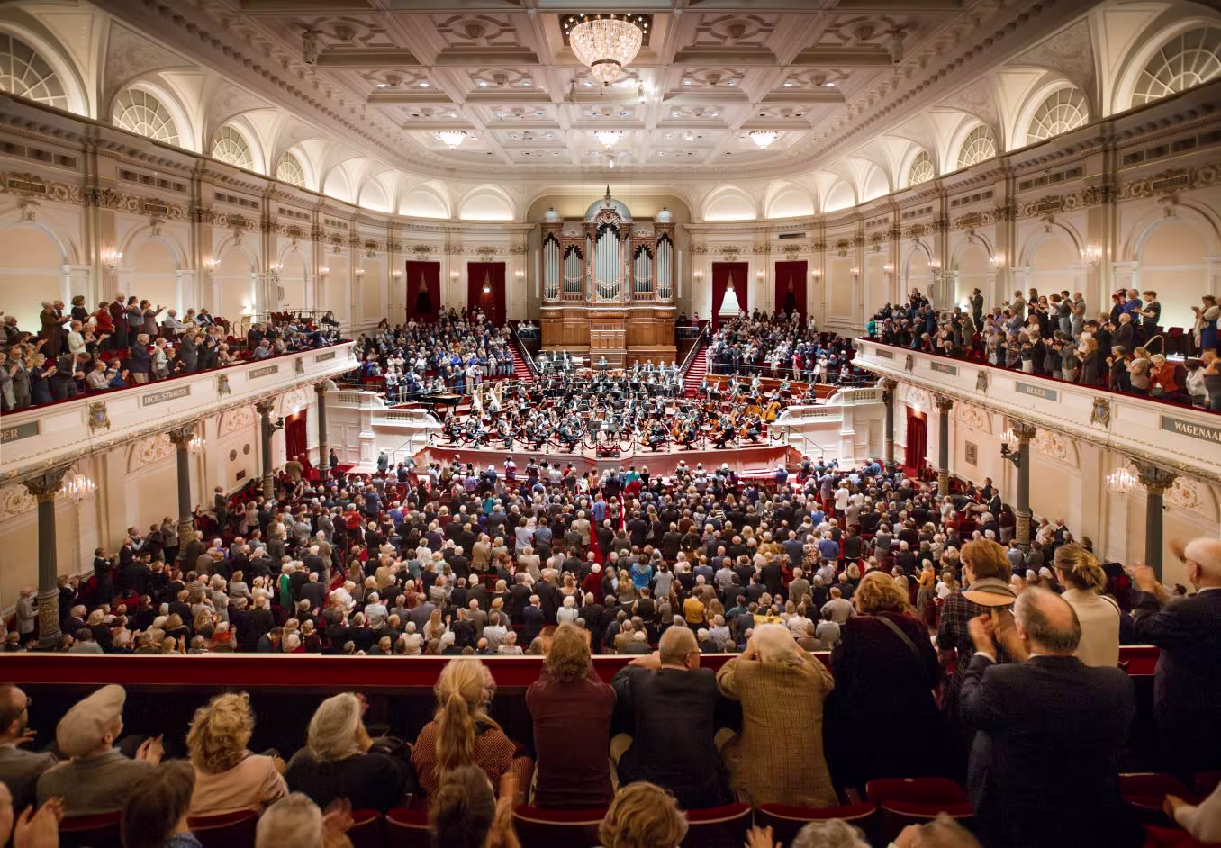 Bomvolle concertzaal met publiek op alle verdiepingen en orkest op het podium in klassieke architectuur.